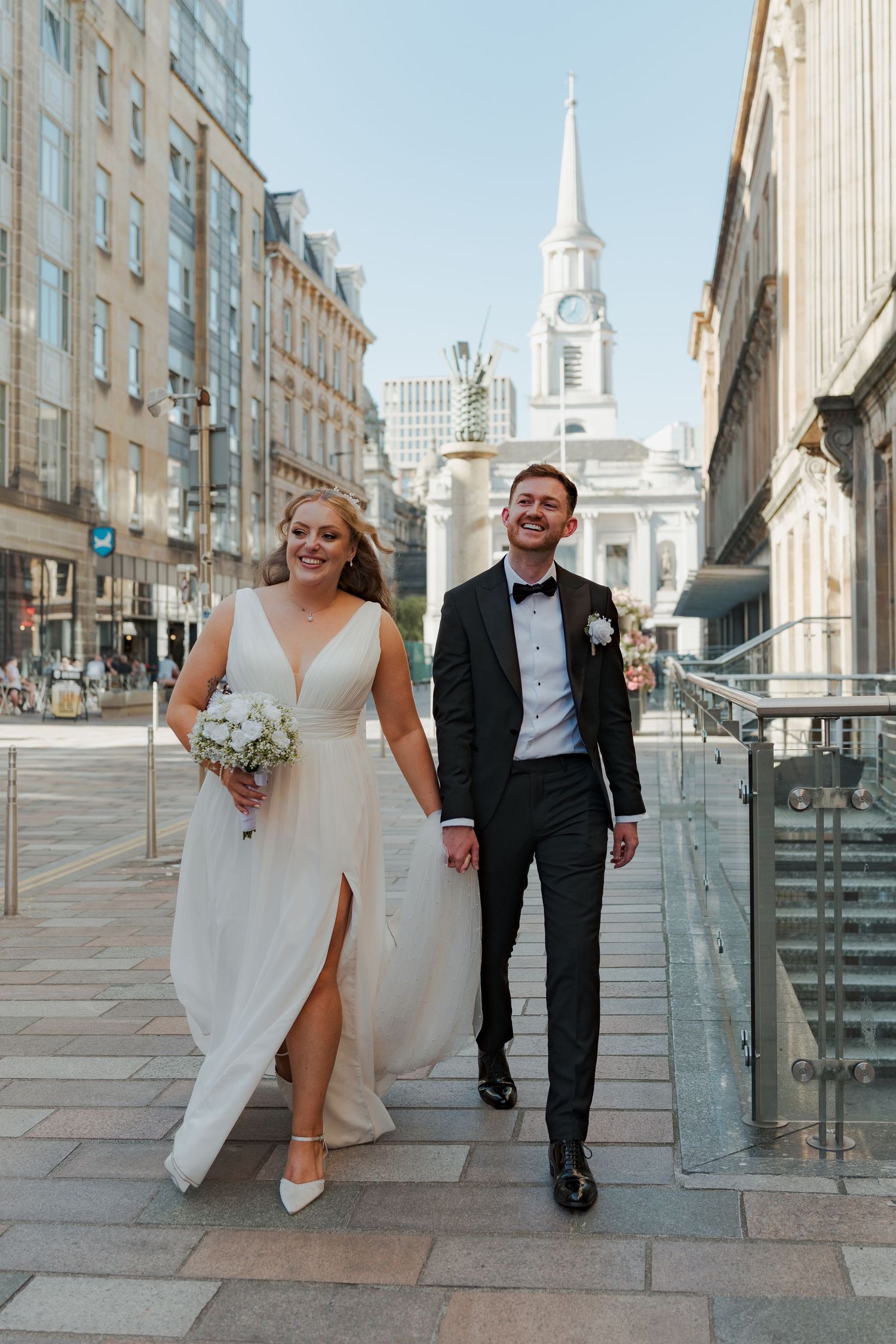 A happy couple in wedding attire walk and laugh together on a city street with a white church steeple visible in the background at Citation.