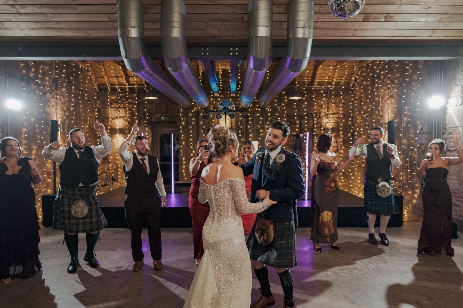 Wedding guests dance and celebrate at Falside Mill decorated with string lights and purple uplighting.