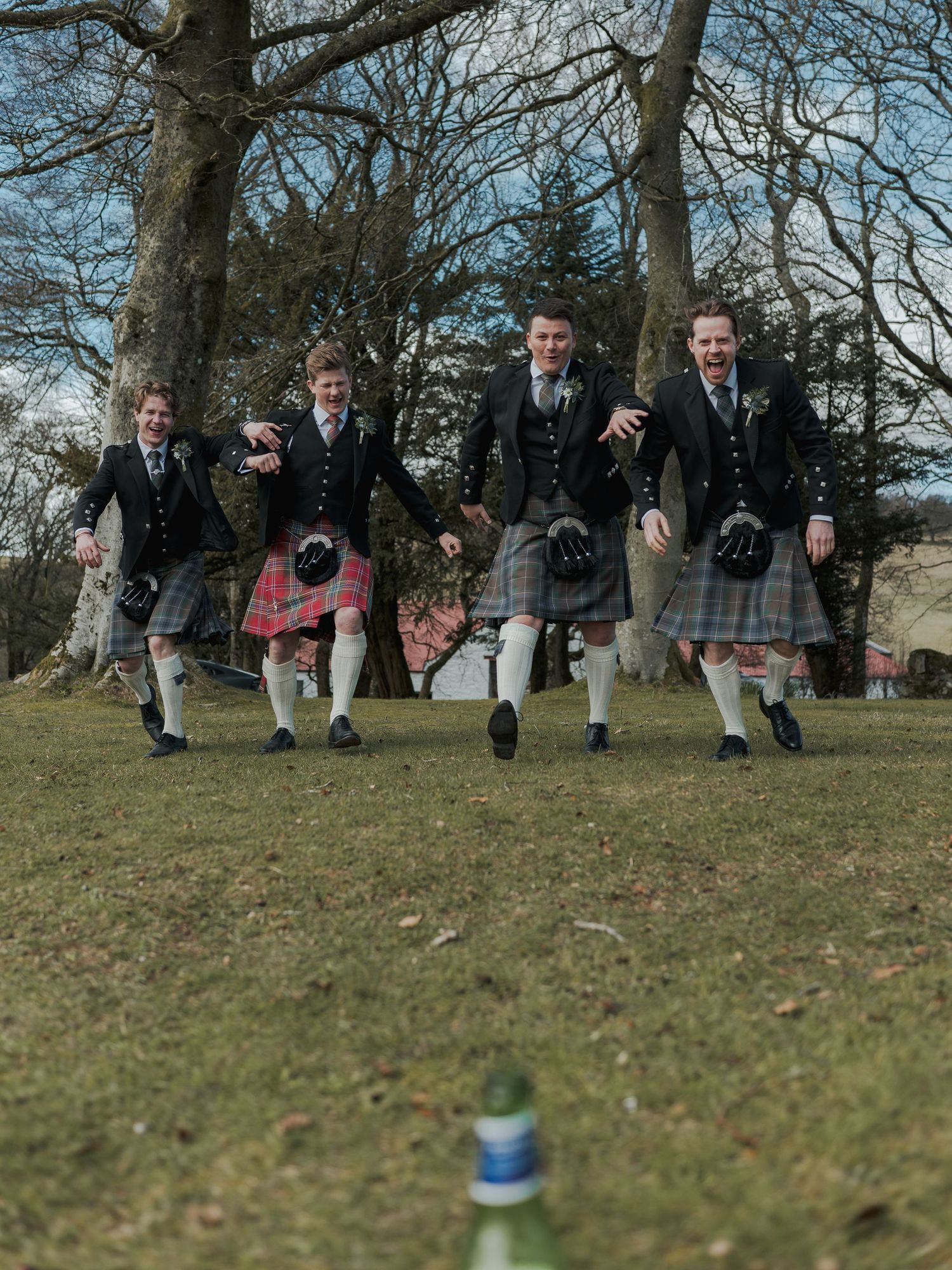 Four men in traditional Scottish kilts and jackets joyfully running across a grassy field with trees in the background.