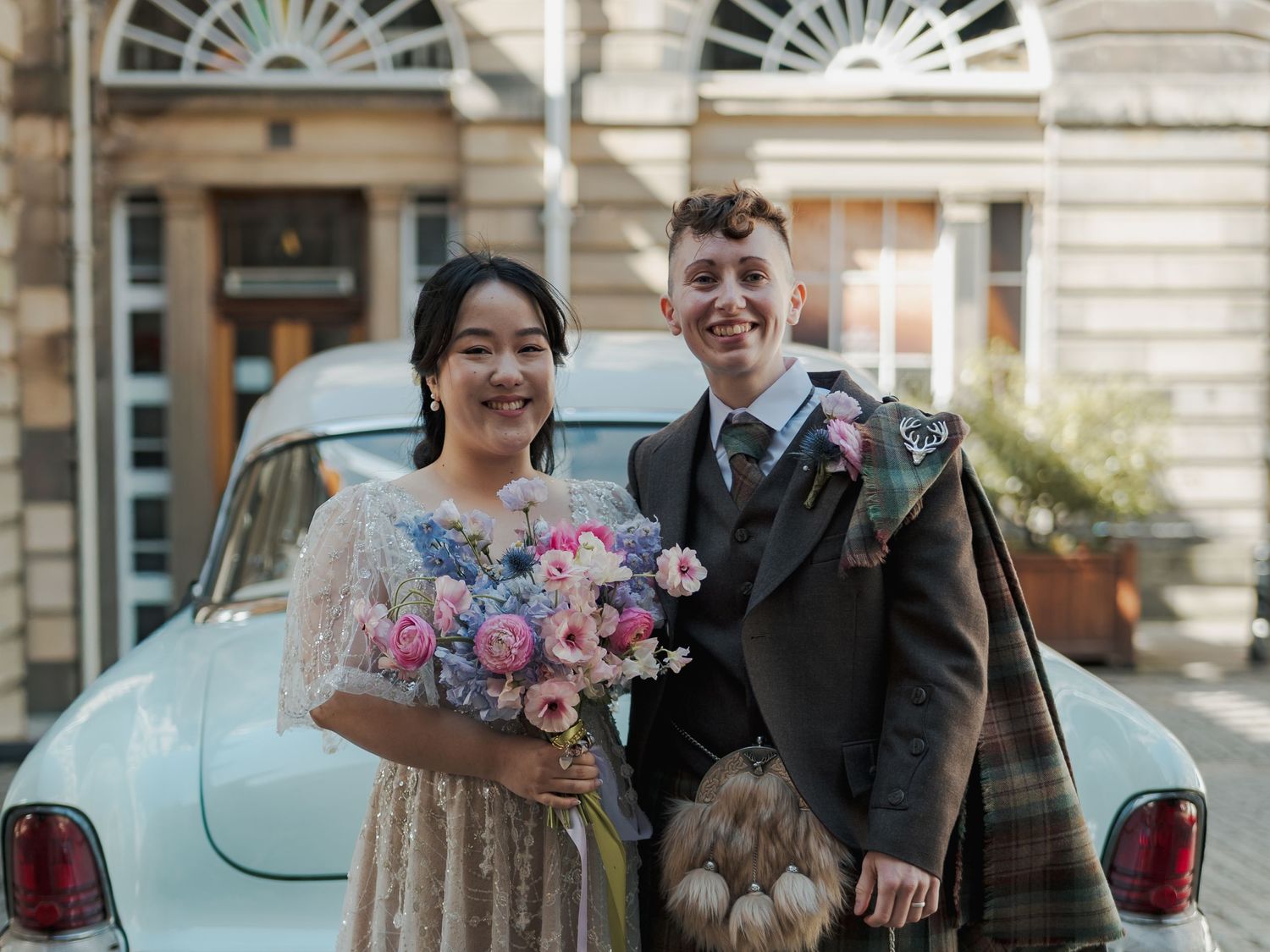 Couple in wedding attire, bride holding pink bouquet, groom in tartan kilt, posing by vintage car on cobblestone street.