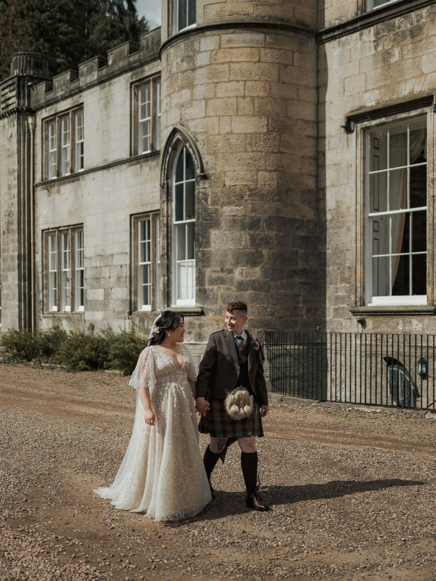 A bride in a white lace gown and groom in a Scottish kilt walk together outside a historic stone castle.