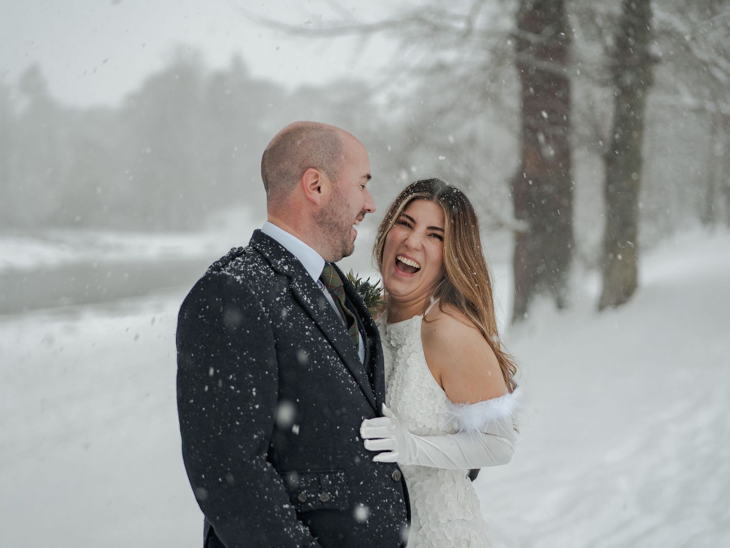 Couple sharing a joyful moment in falling snow, bride in white gown and groom in dark suit, winter wedding portrait.