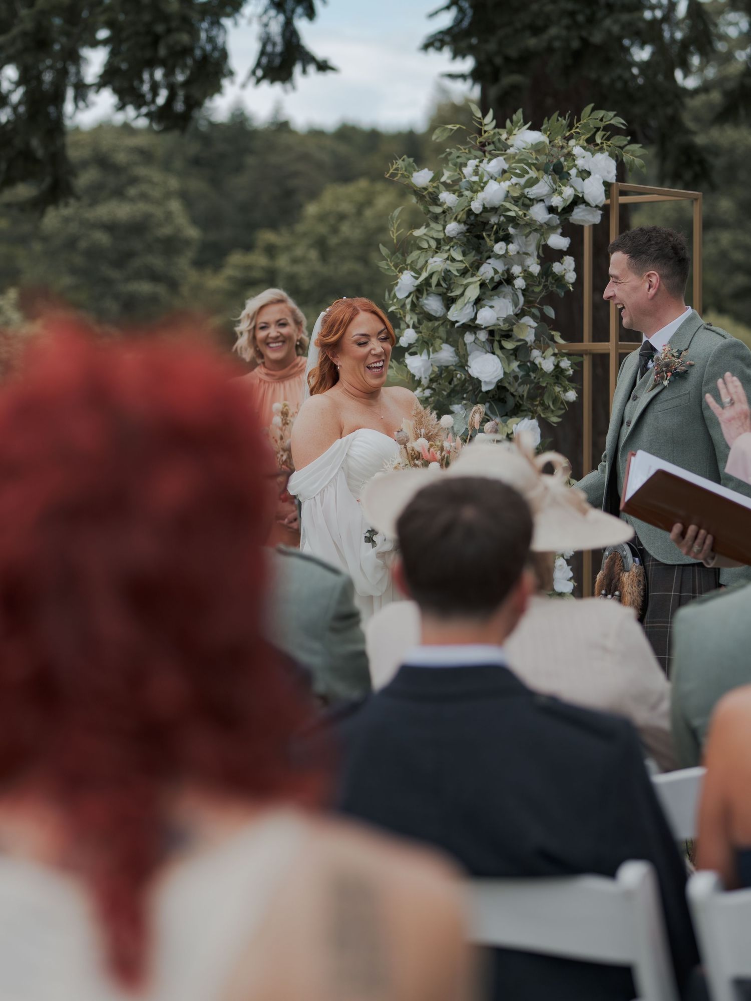 A joyful wedding reception moment captured with guests laughing together under outdoor tree coverage.