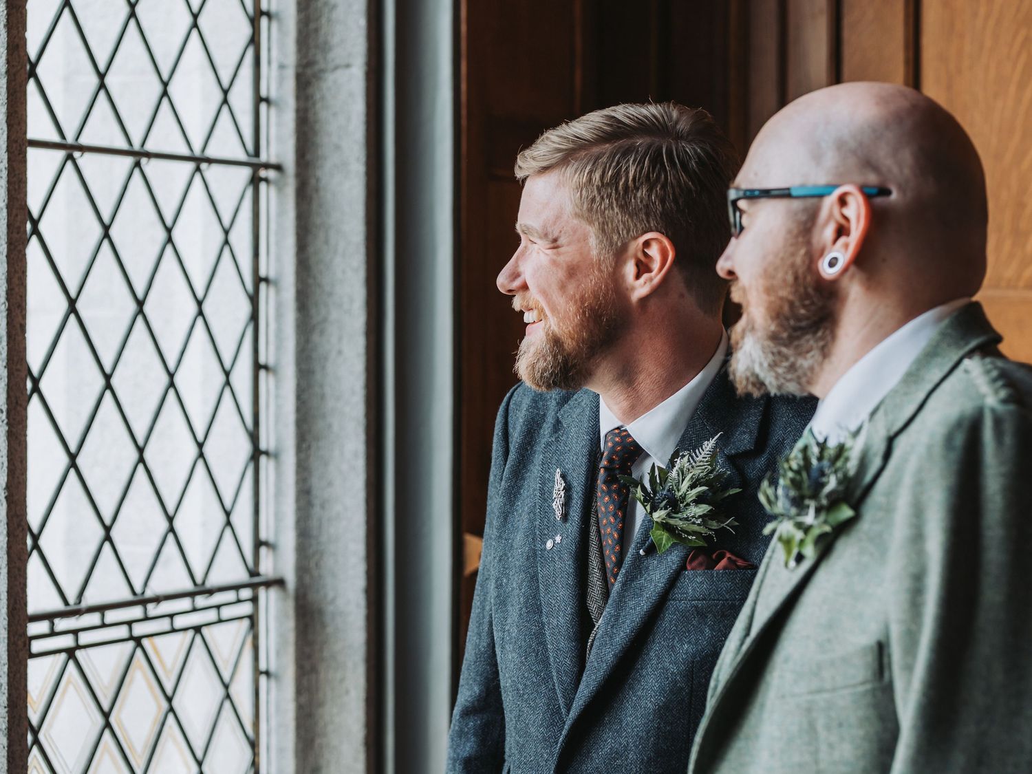 Two groomsmen in dark suits with boutonnieres stand by a gothic window.