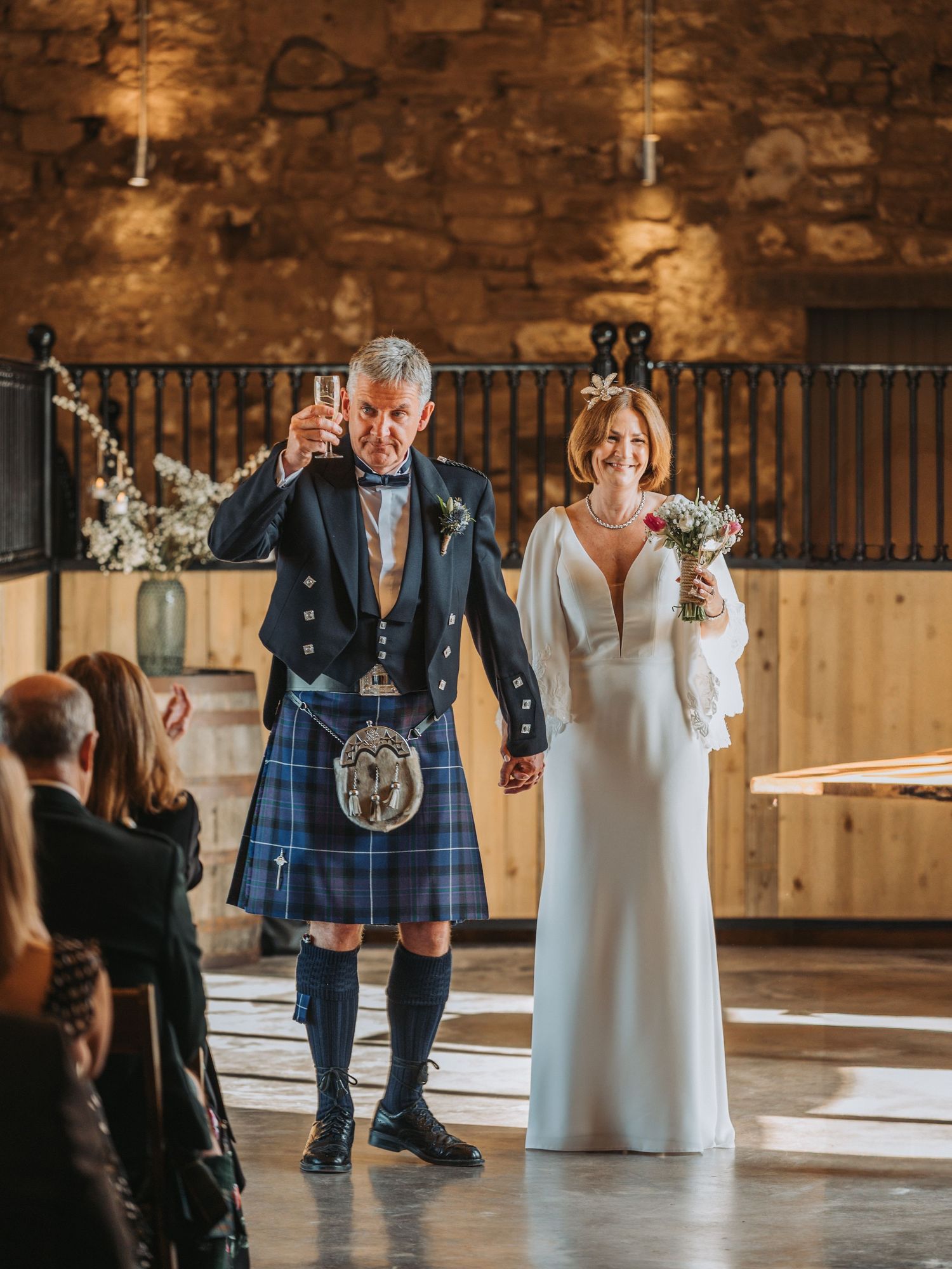 Bride in white dress and groom in traditional Scottish kilt raising hands during wedding ceremony at Falside Mill.