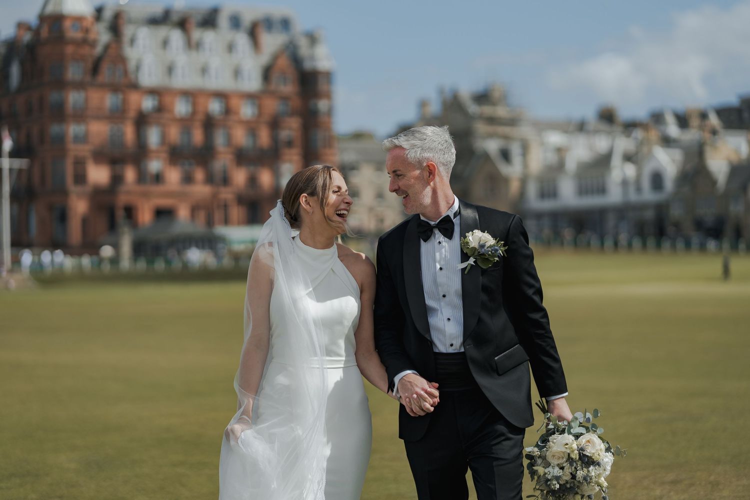 A bride and groom walk hand in hand on a green lawn with a historic red brick building in the background on their wedding day.