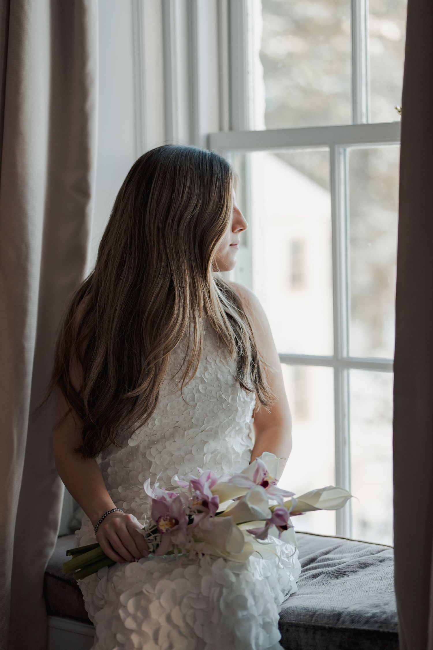 Bride in white lace dress holding pink and white orchid bouquet, seated by a bright window with curtains.
