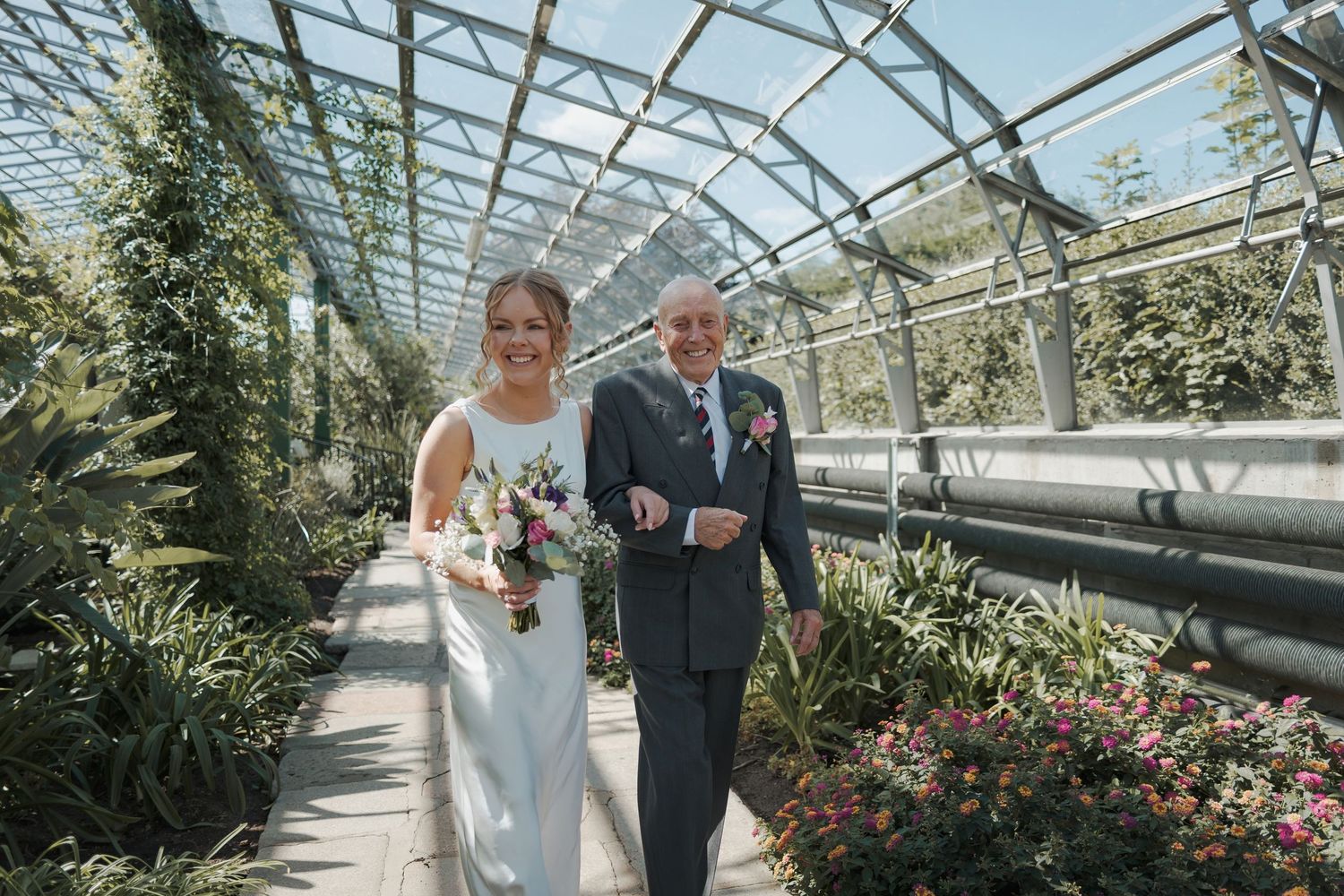 A wedding ceremony takes place inside a beautiful greenhouse filled with lush plants and flowers under a glass roof.
