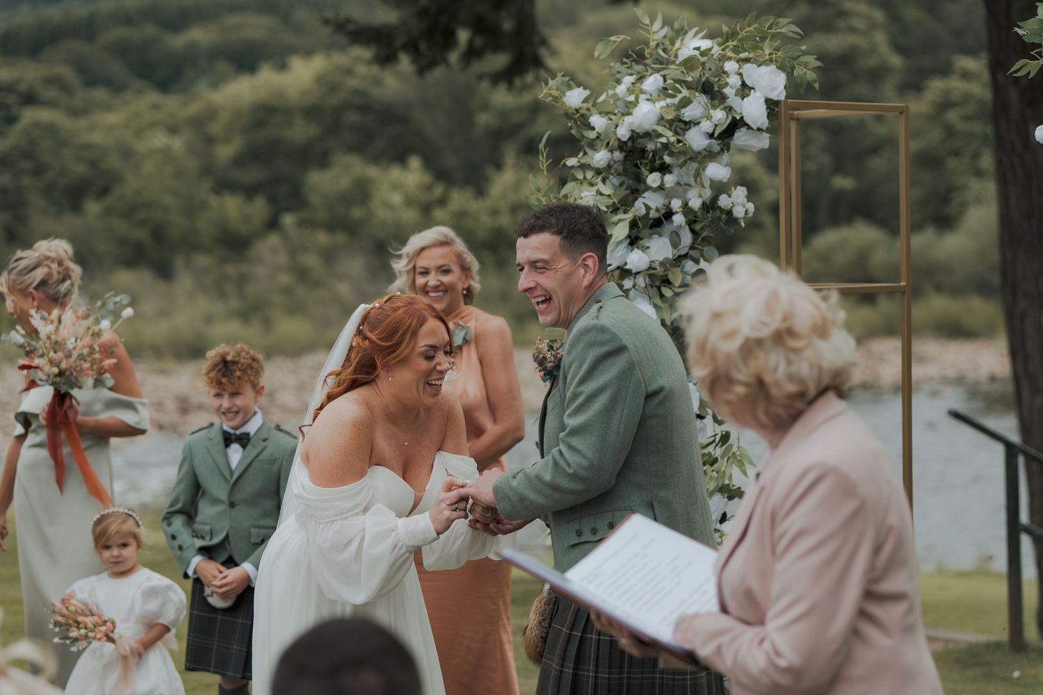 A couple shares a joyful moment during their outdoor wedding ceremony with guests and floral decor in the background.