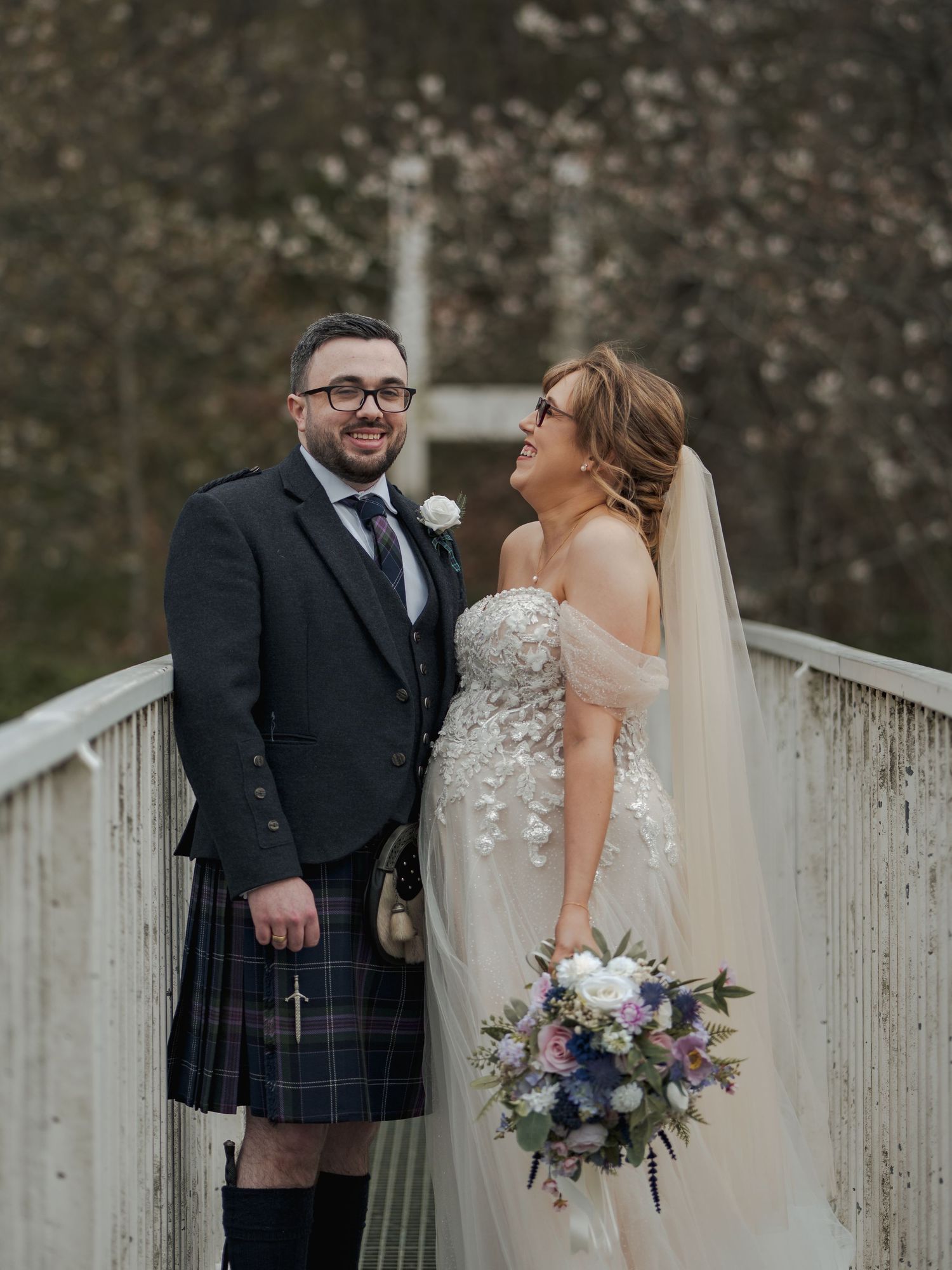 Bride and groom smiling on a bridge, groom in Scottish kilt, bride in floral gown holding a pastel bouquet.