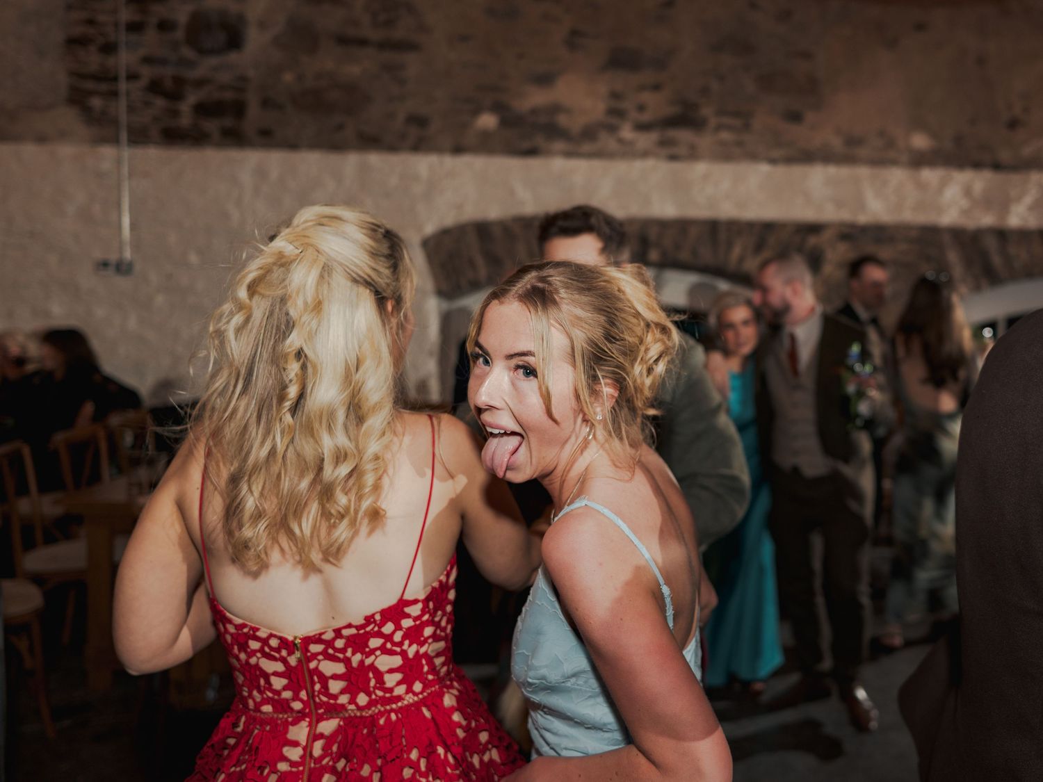 Two young women dancing at a wedding reception, one in red lace dress, other in blue making a fun face.