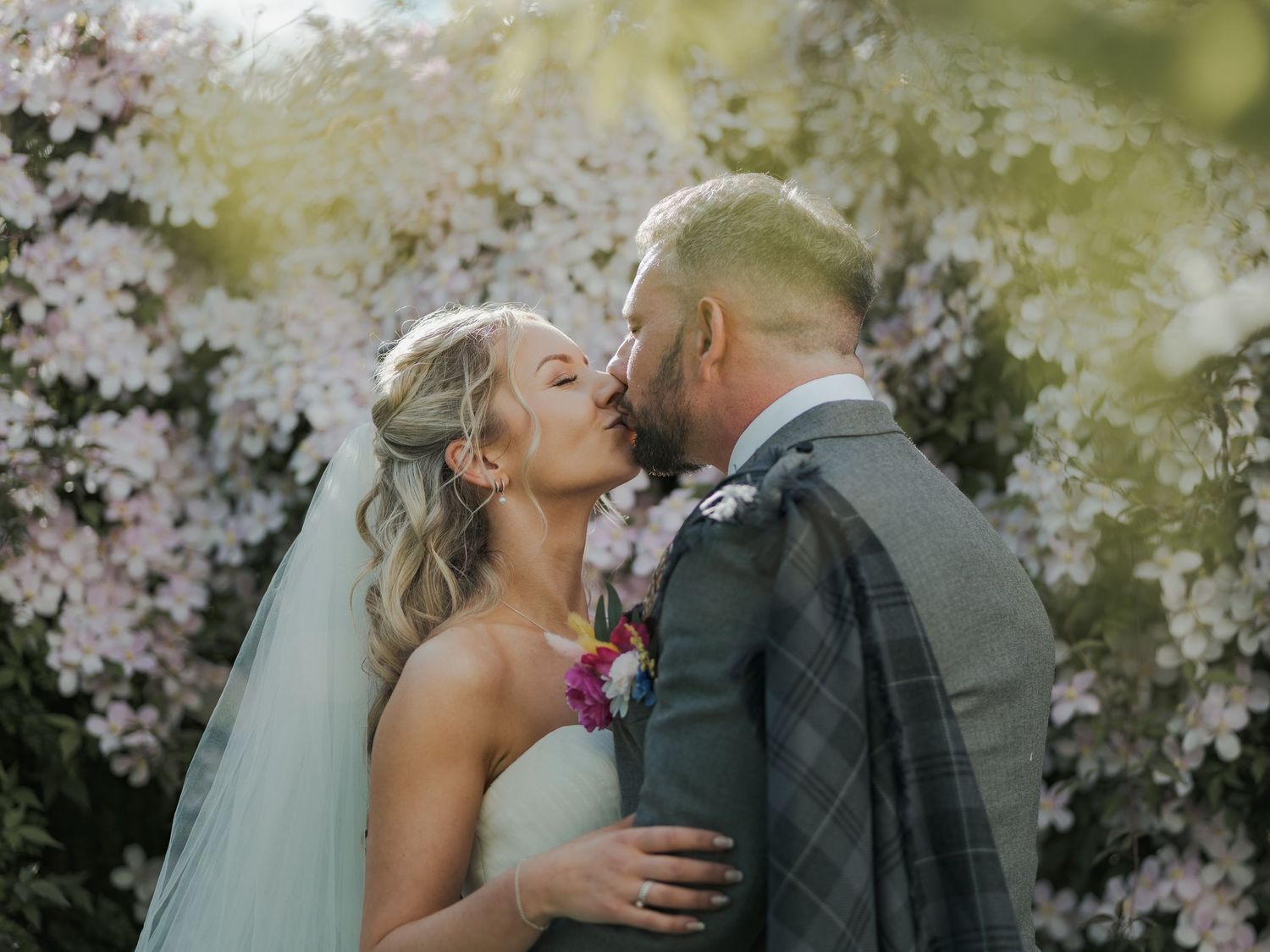 A bride and groom share a romantic kiss surrounded by blooming pink flowers in a lush garden wedding setting.