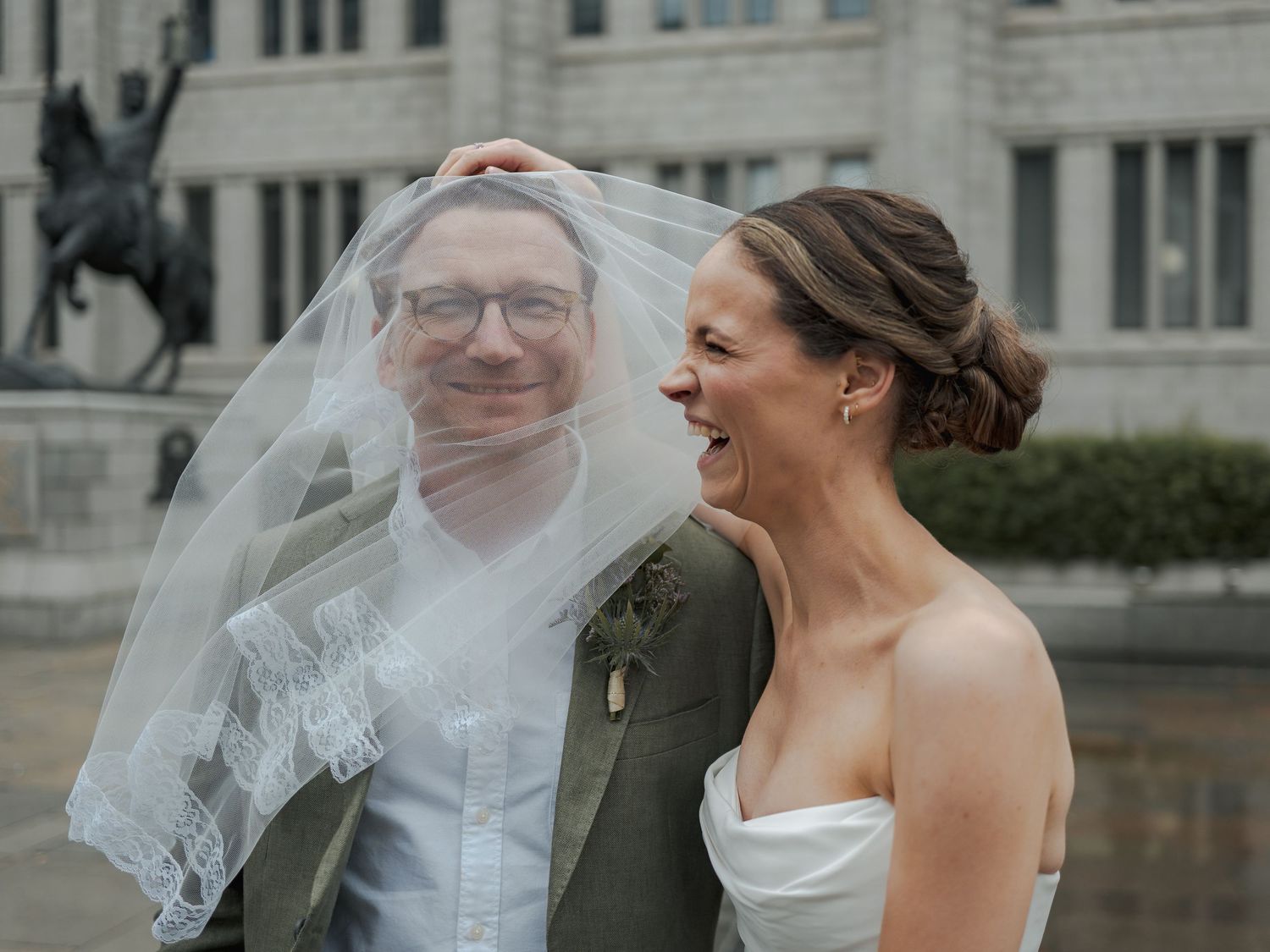 Bride and groom laughing together outdoors, veil blowing in wind, classic building and statue in background.