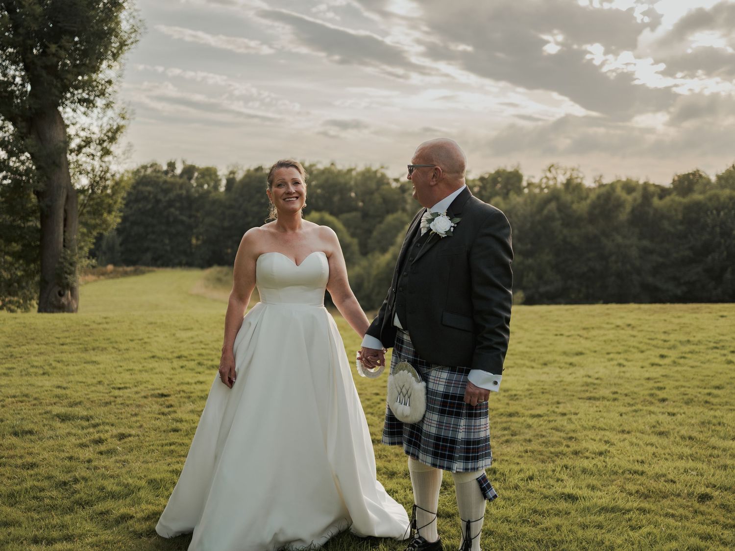 Bride in white gown and groom in blue tartan kilt hold hands on green lawn at outdoor wedding ceremony.