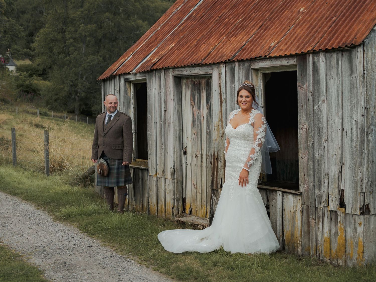 Bride in white lace gown and groom in kilt stand outside a rustic wooden barn on a green hillside.