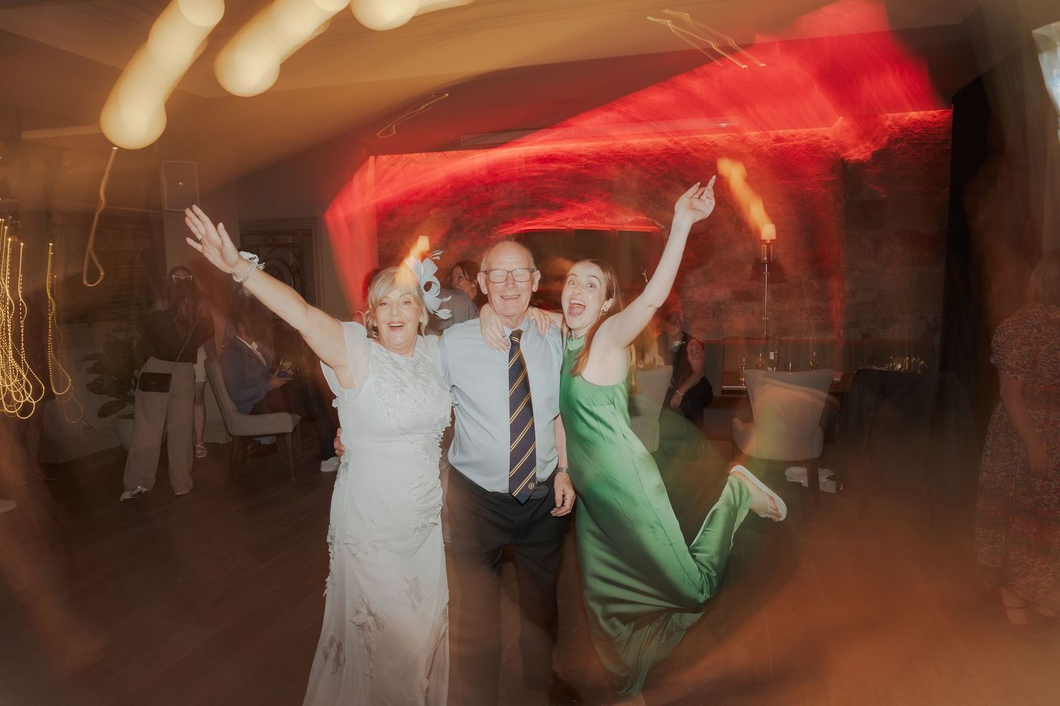 Wedding reception guests dance and celebrate with colorful red lighting creating a festive atmosphere on the dance floor.
