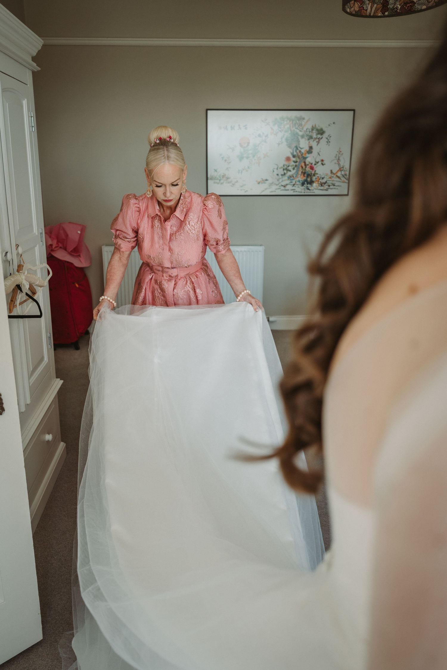 Woman in pink dress helps prepare a flowing white wedding gown in an elegant bedroom setting at Kinclune House.