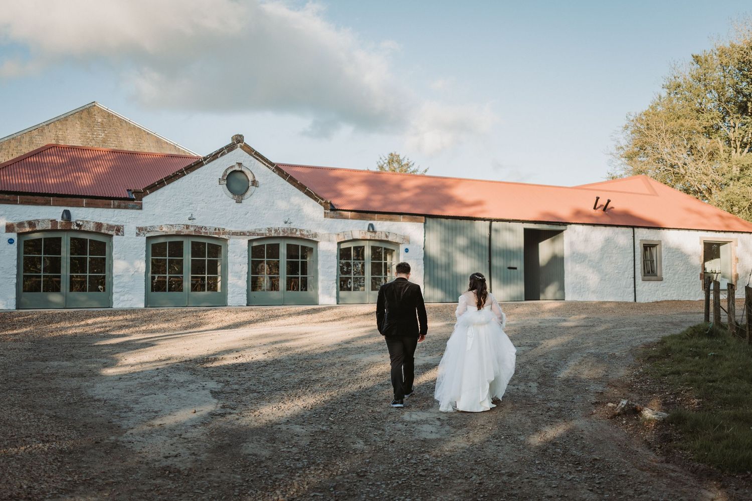 A couple in wedding attire walking on a gravel path beside a Kinclune Steading at sunset.