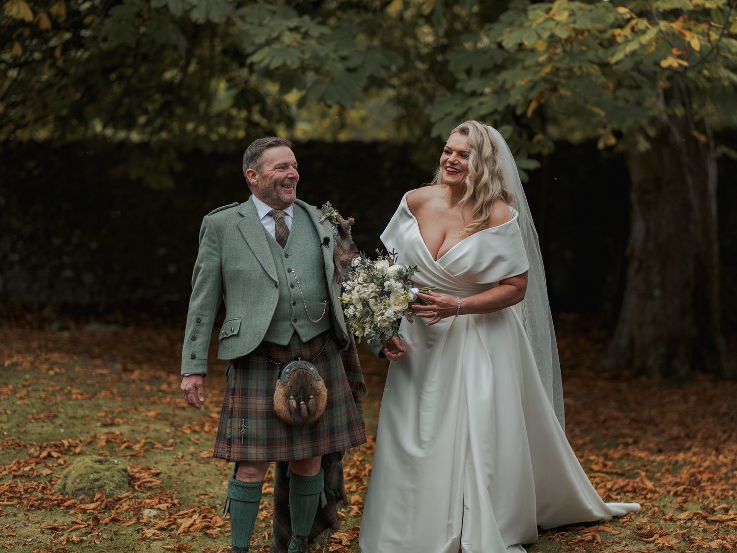 Bride in white gown holding bouquet laughs with groom in green Scottish kilt among autumn leaves.