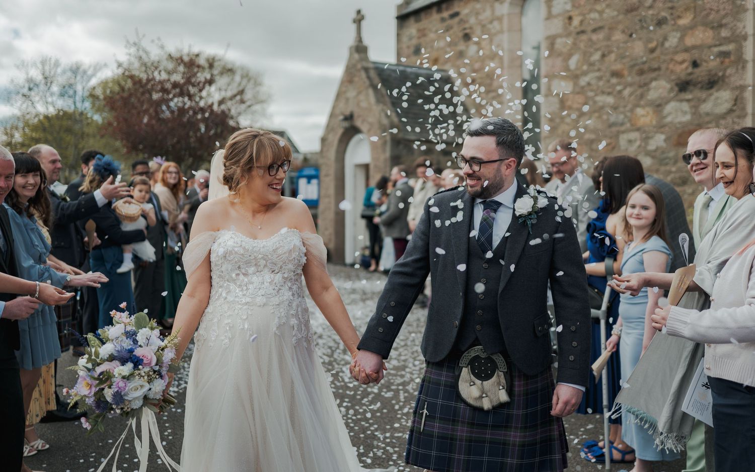 Bride and groom walk hand-in-hand through confetti shower outside stone church, groom wearing traditional Scottish tartan kilt.