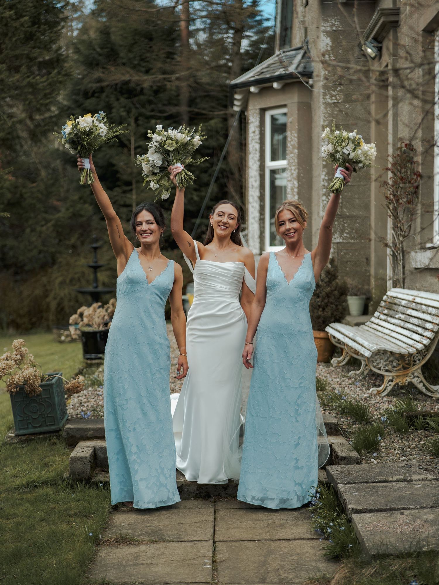 Bride in white gown flanked by two bridesmaids in blue dresses holding bouquets aloft outside a stone manor.