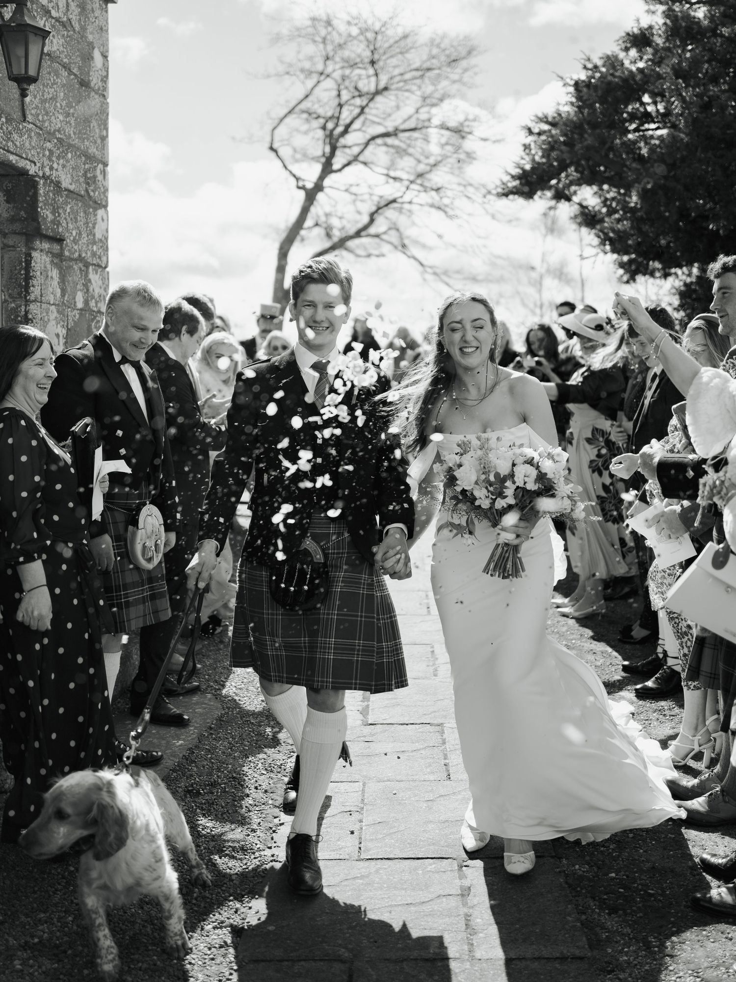 Black and white photo of a smiling bride and groom in tartan kilt walking through confetti with guests and a dog.