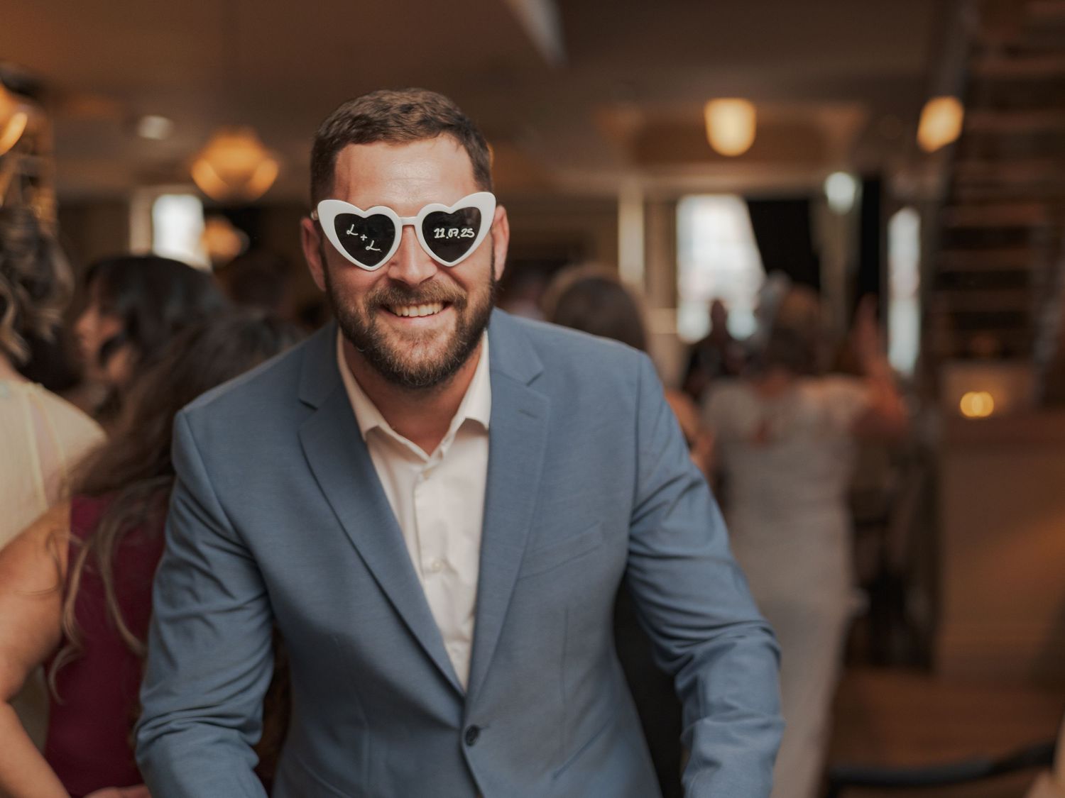 A guest wearing heart-shaped sunglasses and a blue blazer smiles at a wedding reception.