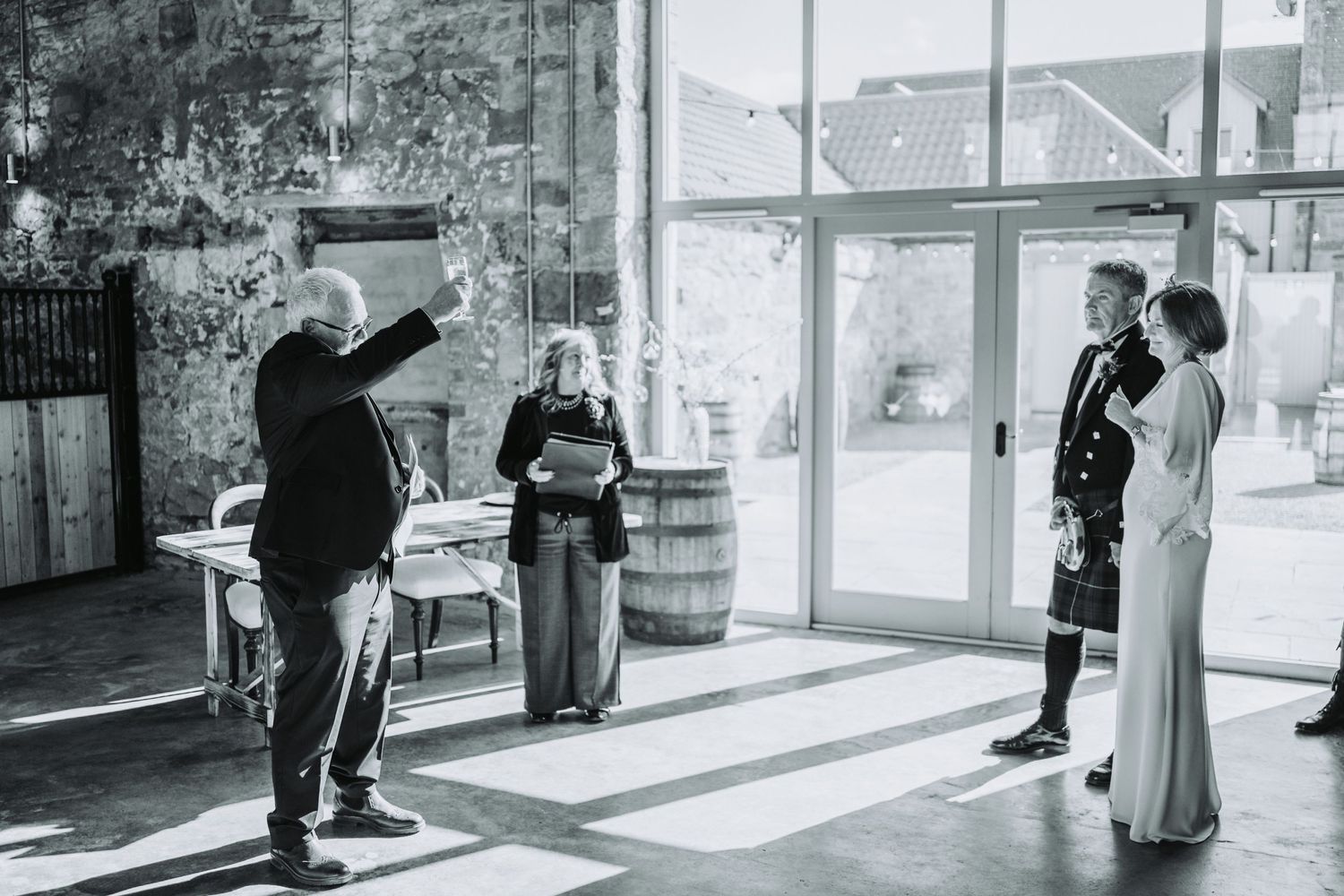 Black and white photo of wedding officiator gesturing during ceremony in sunlit Falside Mill.