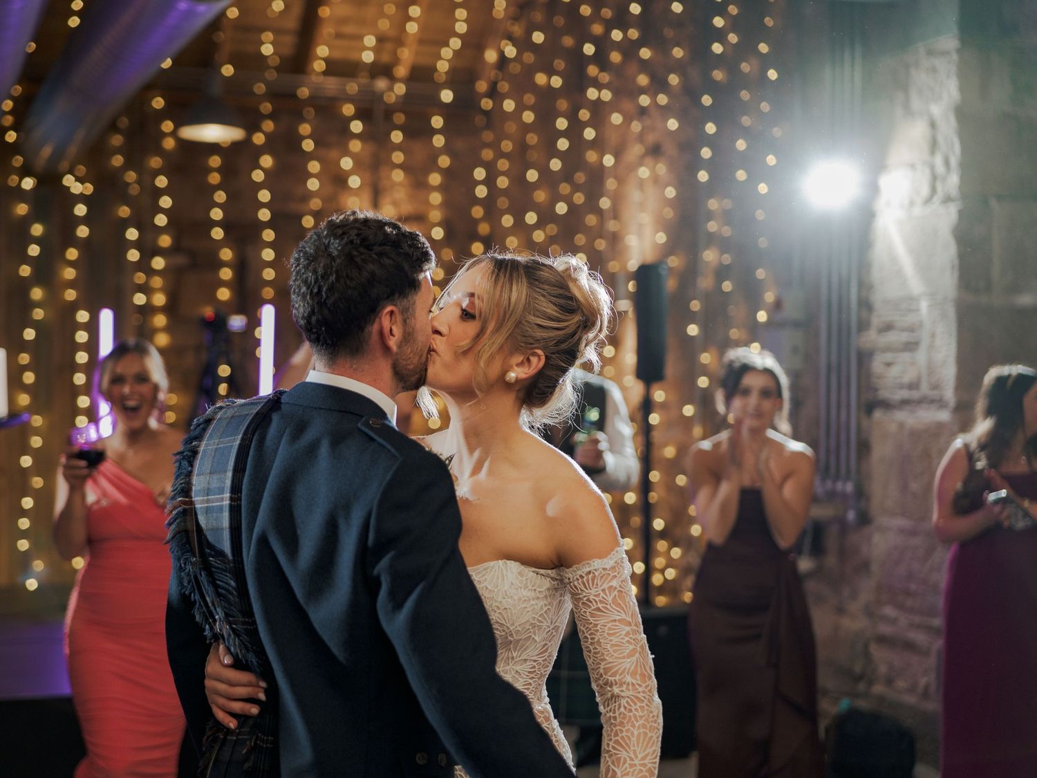 A couple shares an intimate kiss on their wedding day at Falside Mill surrounded by twinkling lights and guests in a romantic venue.