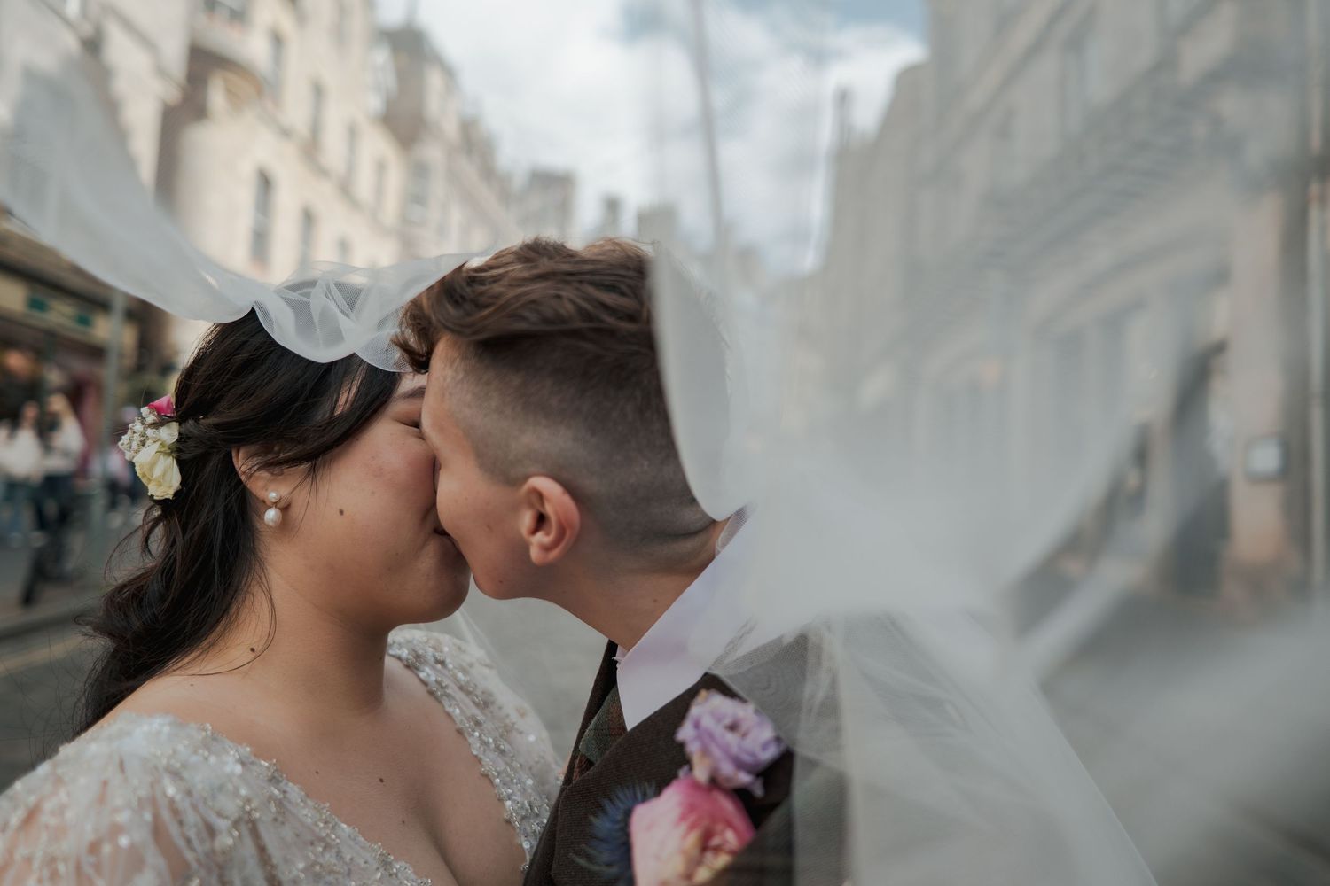 Bride and groom sharing a romantic kiss under a flowing veil on a city street on their wedding day.