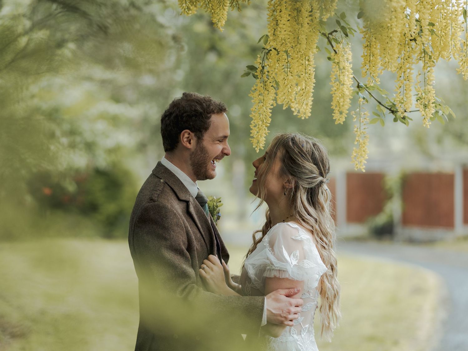 A smiling bride and groom share a romantic moment under yellow laburnum flowers in a lush garden setting.