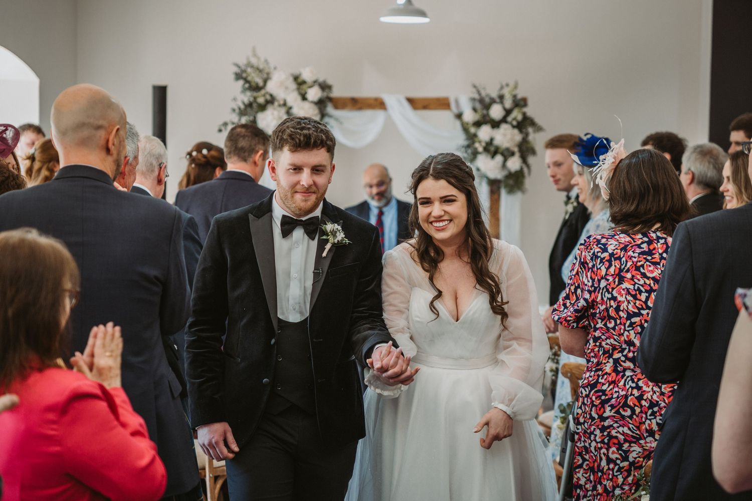 A joyful couple in wedding attire walk together through a crowd of guests at the indoor reception venue, Kinclune Steading.