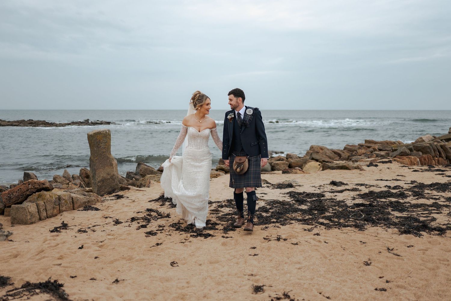 A couple in wedding attire stands on a rocky beach coastline on an overcast day in Scotland.