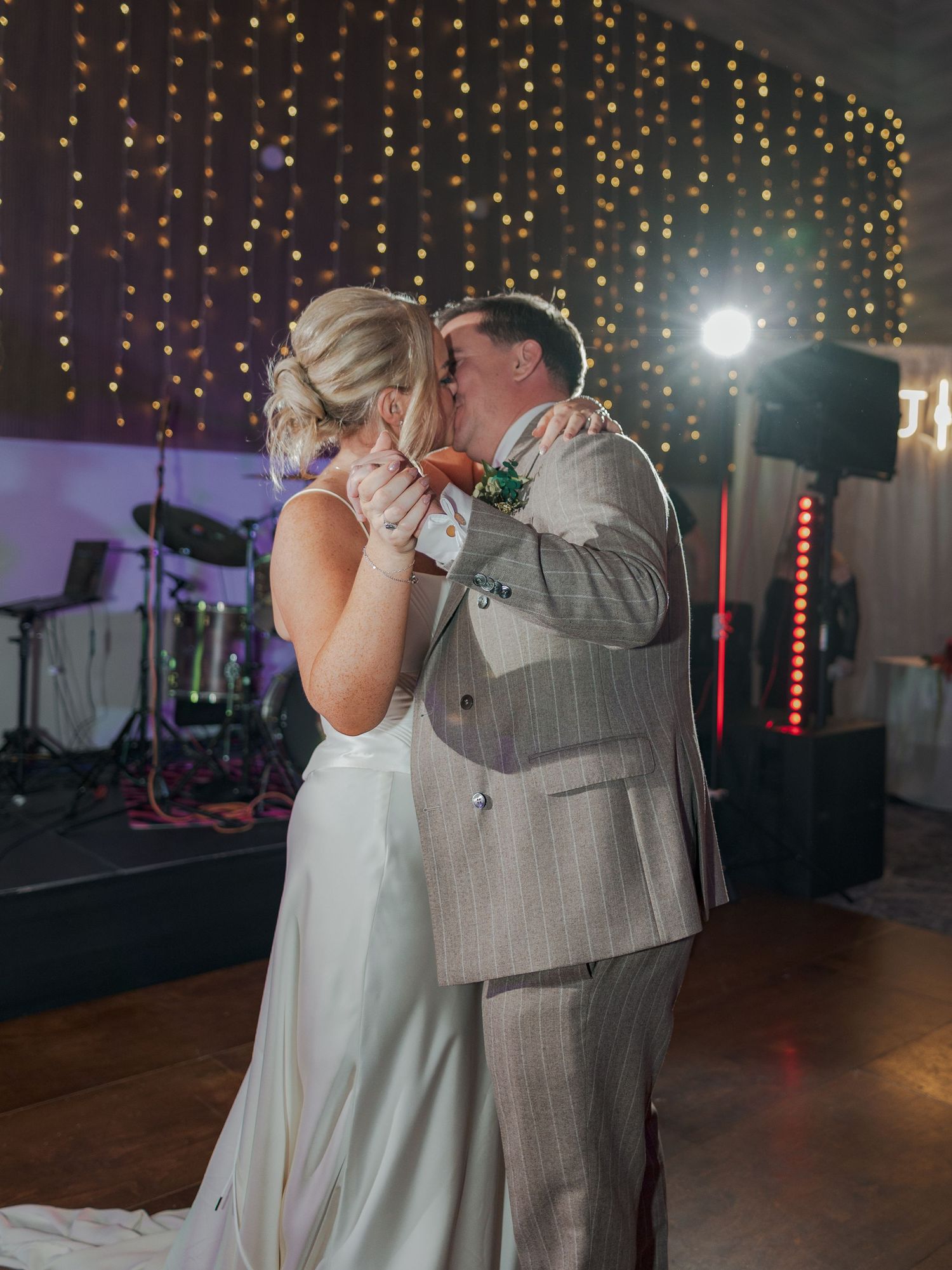 A couple shares a romantic dance moment under twinkling string lights at an indoor wedding reception.