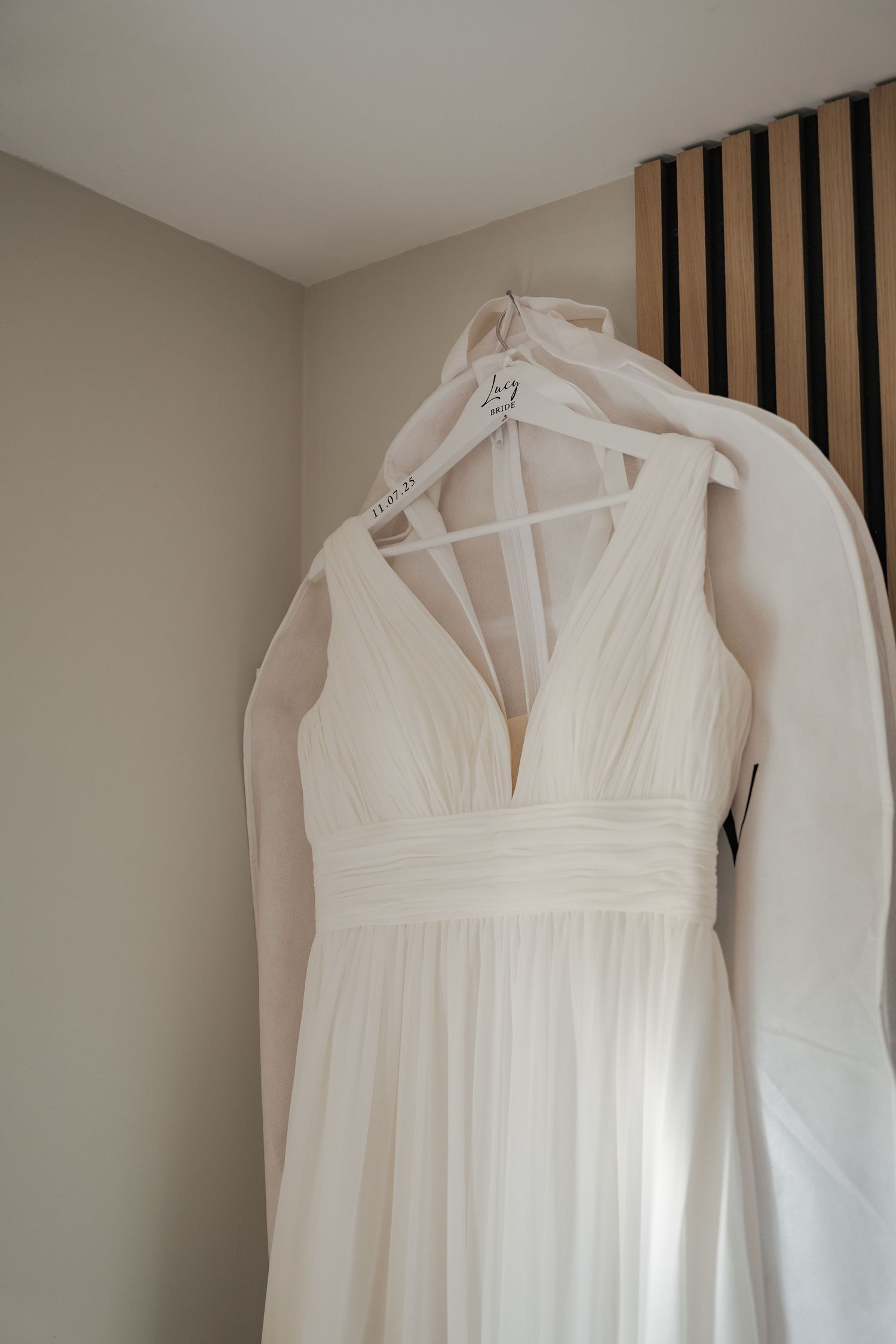 A white halter wedding dress with a plunging neckline hangs elegantly against a neutral wall with vertical wooden slats.