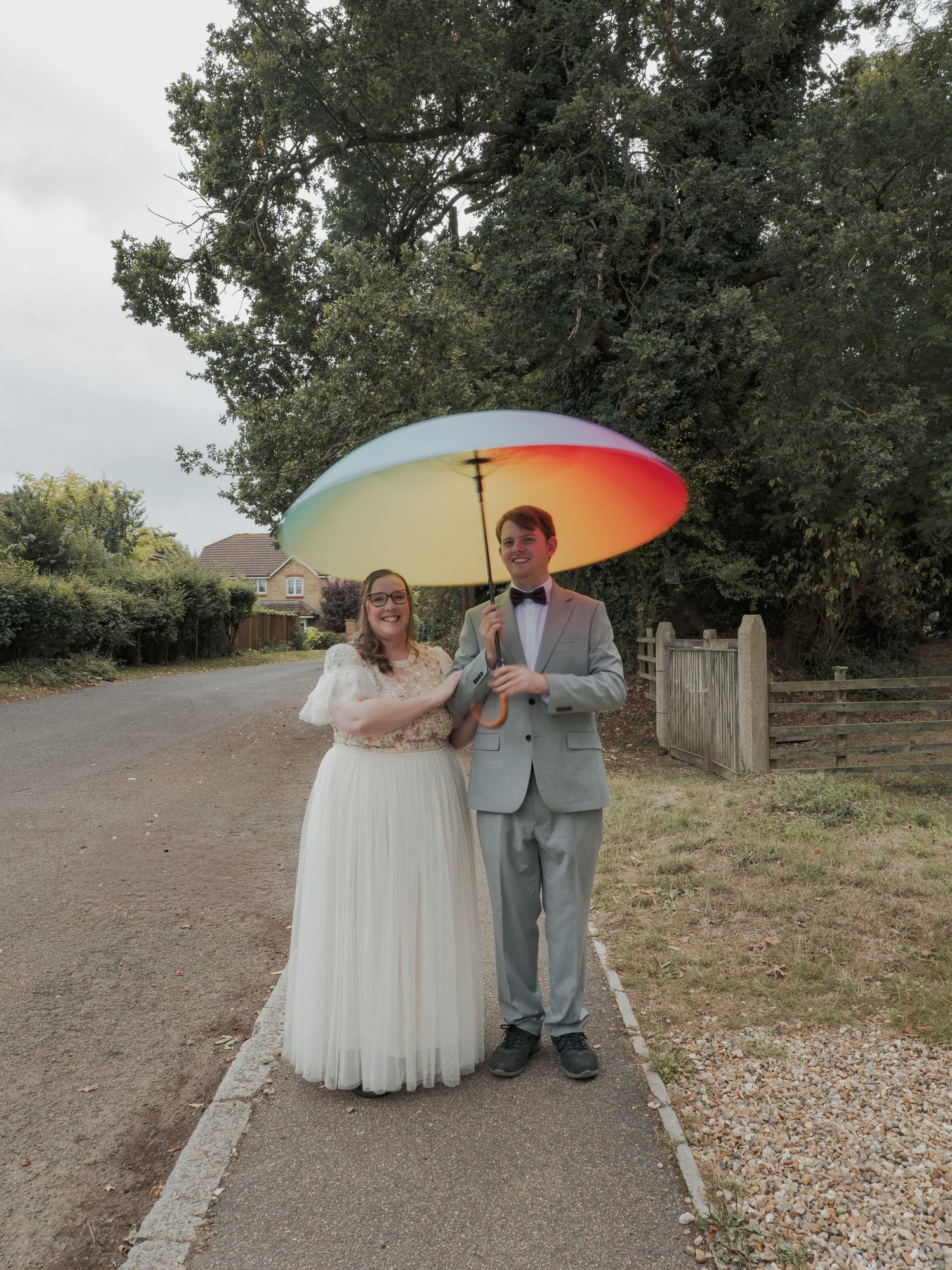 A wedding couple poses under a rainbow umbrella against a cloudy sky on a rocky gravel path beside green trees.