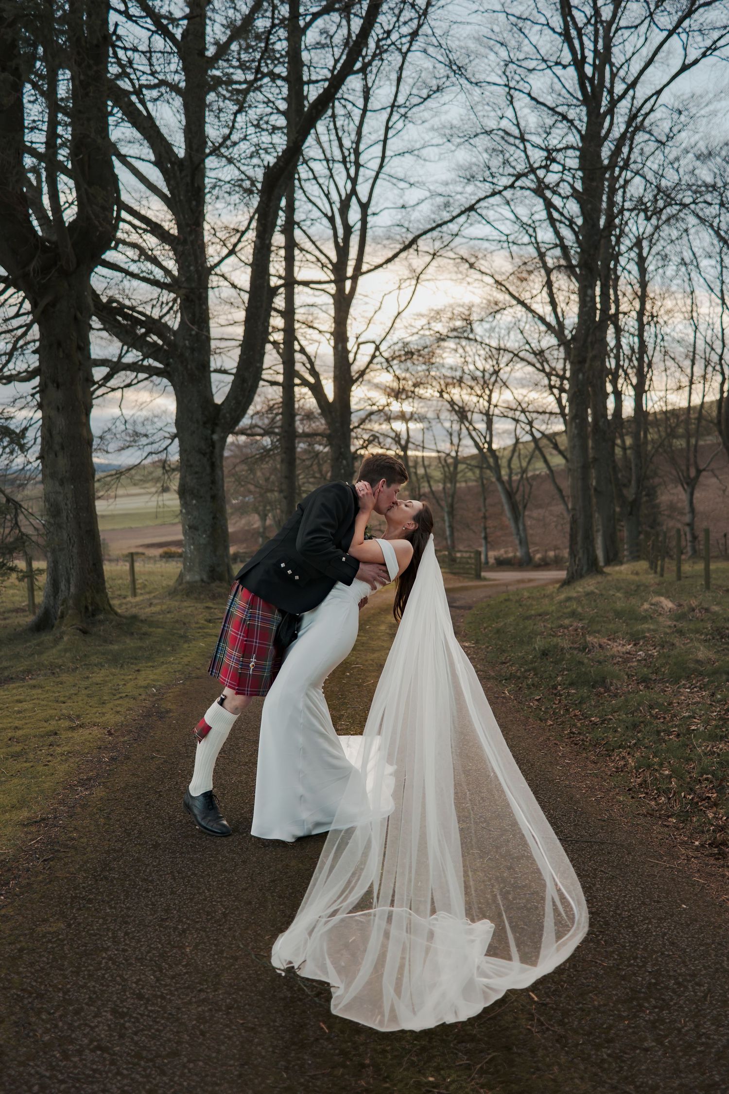 Bride and groom share a romantic dip kiss on a tree-lined path at sunset, groom in tartan kilt, bride in flowing gown with veil.