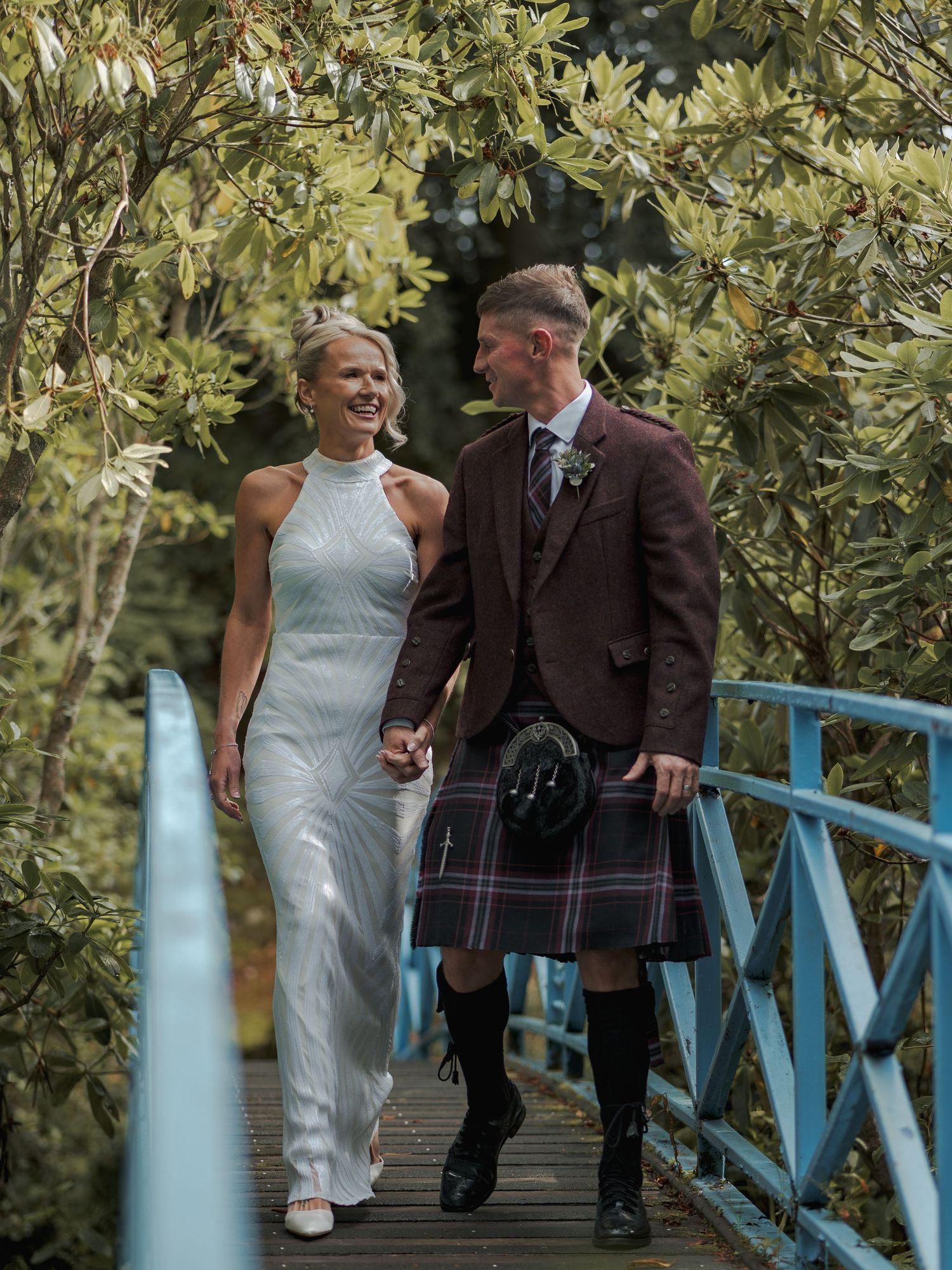 Bride in white halter gown and groom in tartan kilt walk hand-in-hand on blue bridge outdoors.