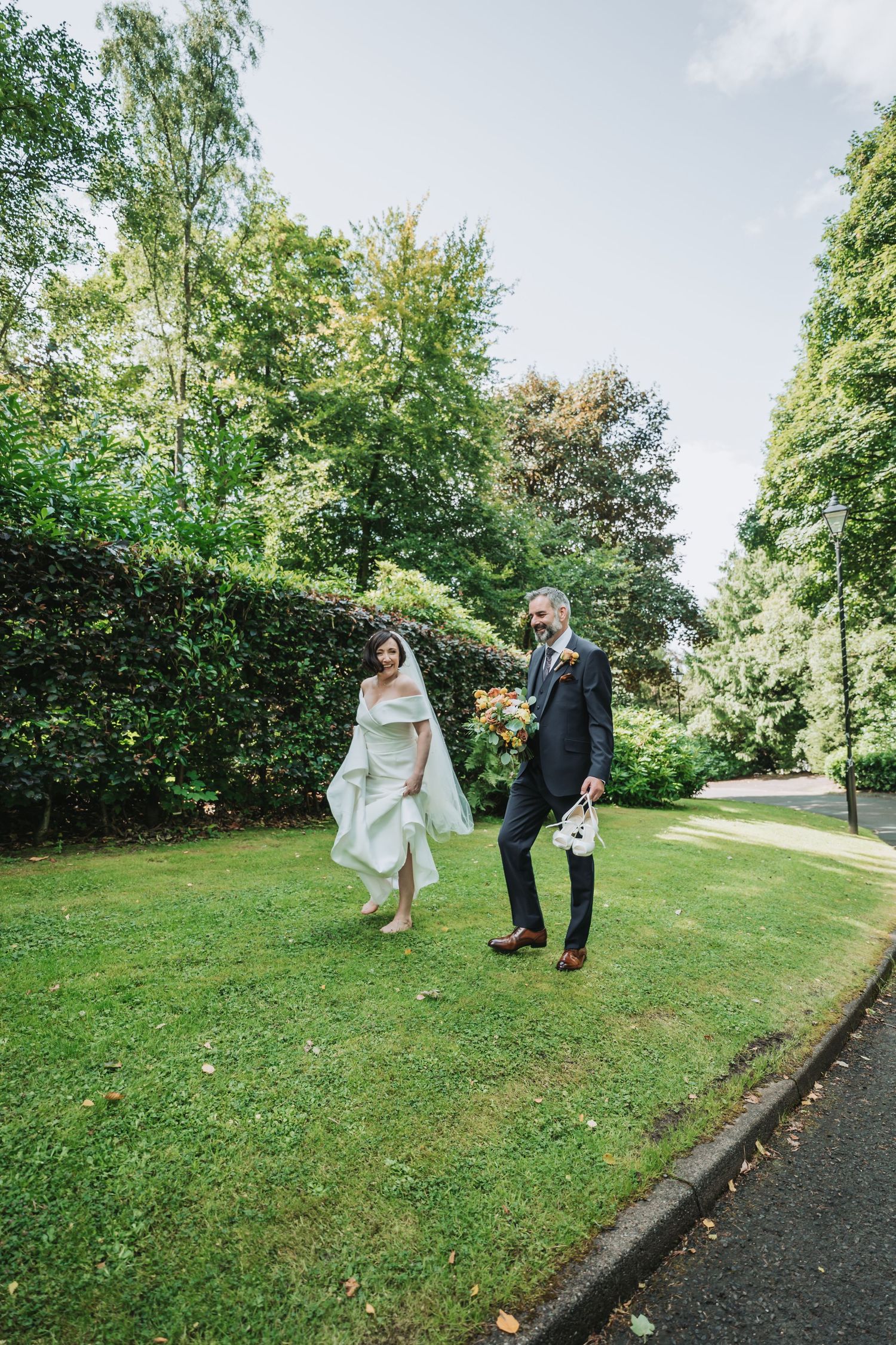 A couple strolls hand in hand on a lush green lawn of Dunblane Hydro surrounded by tall trees and hedges on a bright summer day.