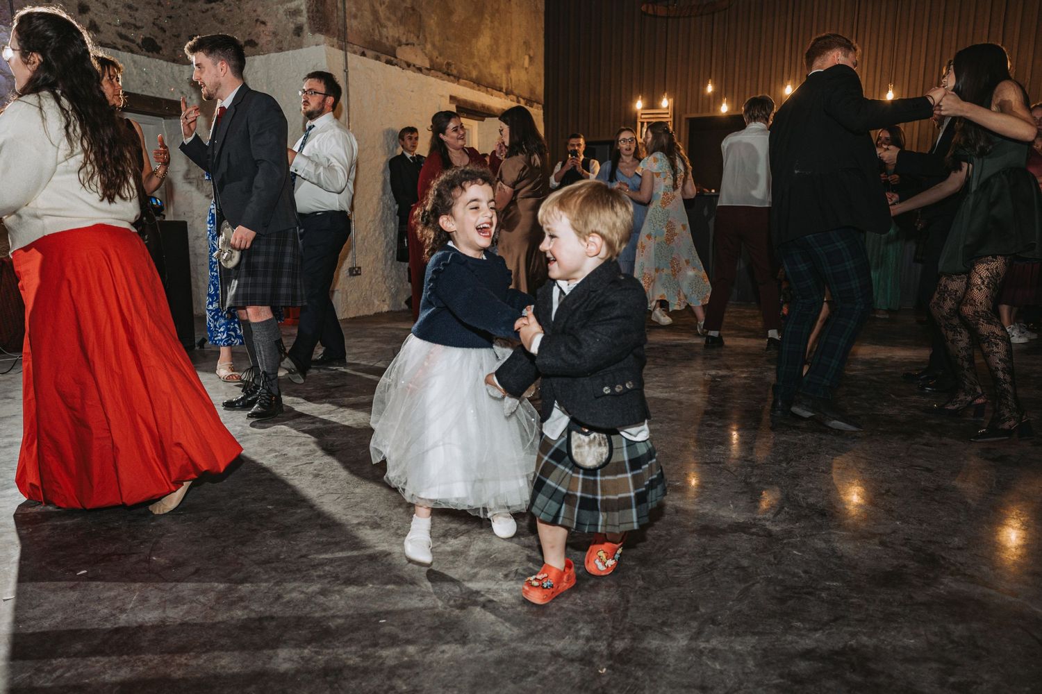 Two young children in formal attire dance together at a wedding reception on a dark wooden floor.