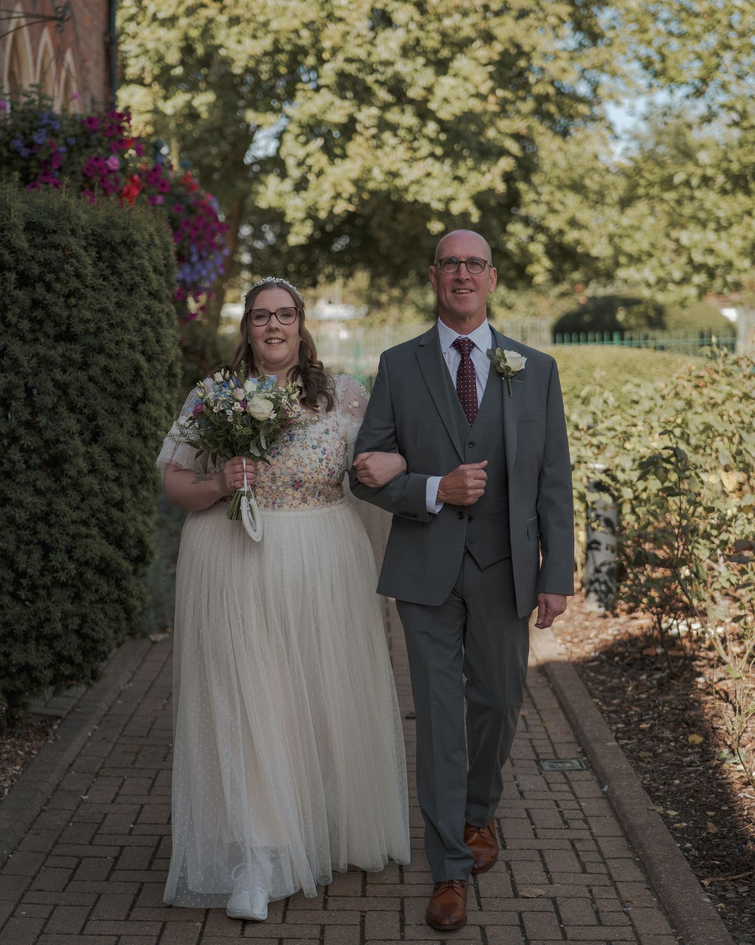 Two people walk arm in arm down a brick pathway surrounded by lush greenery and blooming flowers on a sunny day.