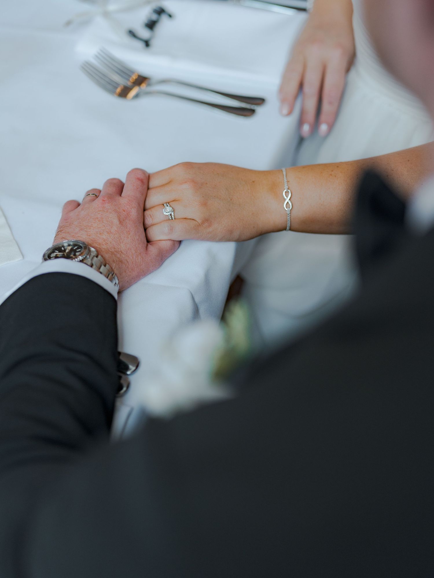Close-up of hands touching on a white tablecloth with wedding rings and formal attire visible.