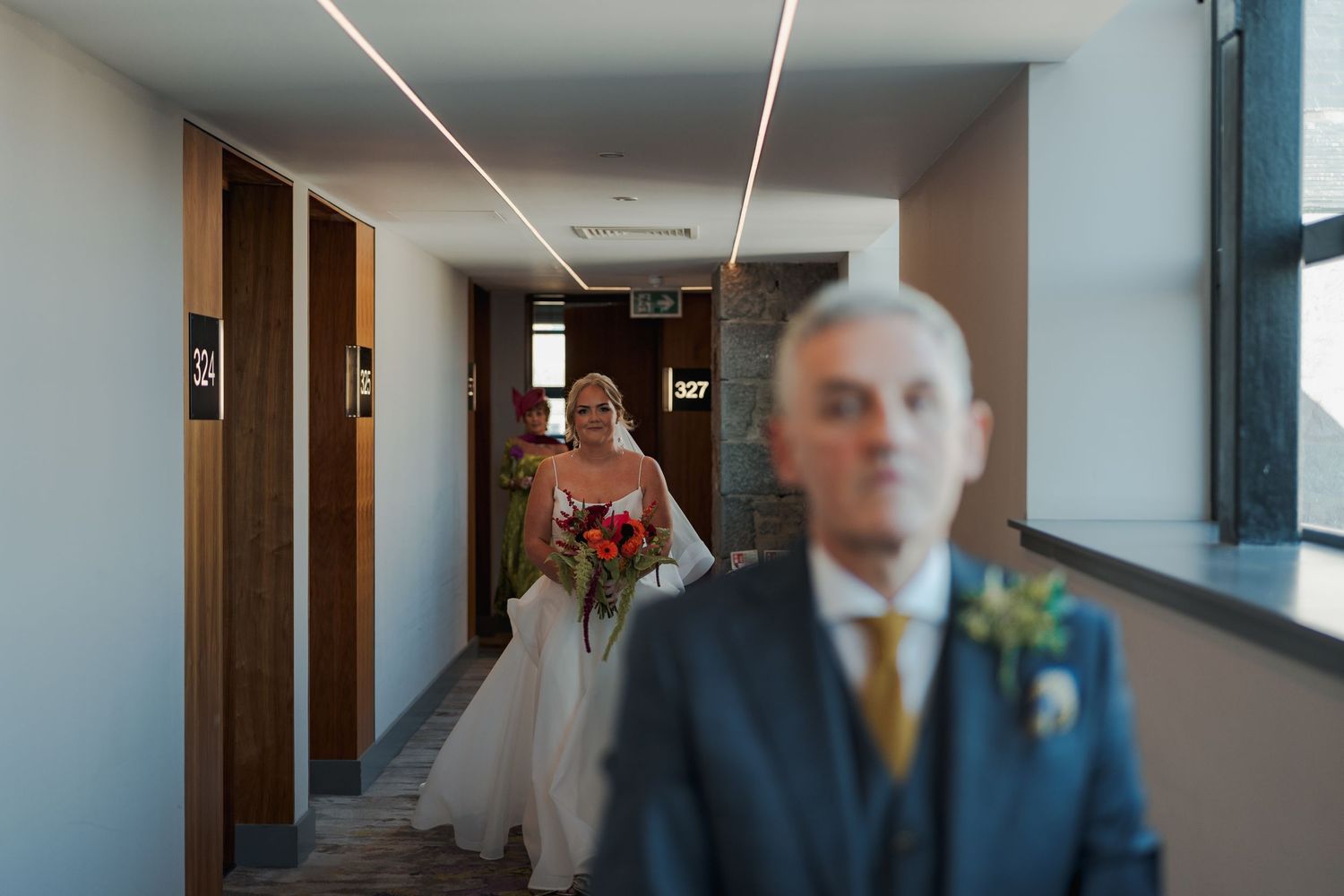 A bride in a white wedding dress walks down a modern hotel hallway with natural light streaming through windows.