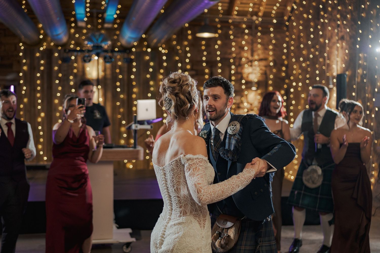 Wedding guests dancing together at Falside Mill under twinkling string lights at an indoor reception venue.