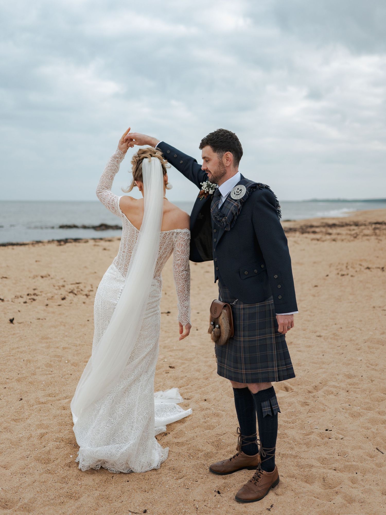A couple dancing playfully on a cloudy beach, with the groom wearing a traditional Scottish kilt and the bride in white.