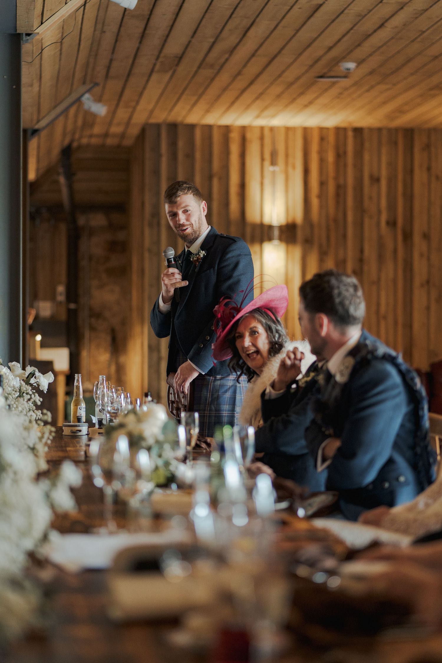 Wedding guests share laughter and conversation during a reception in a rustic wooden venue with warm lighting.