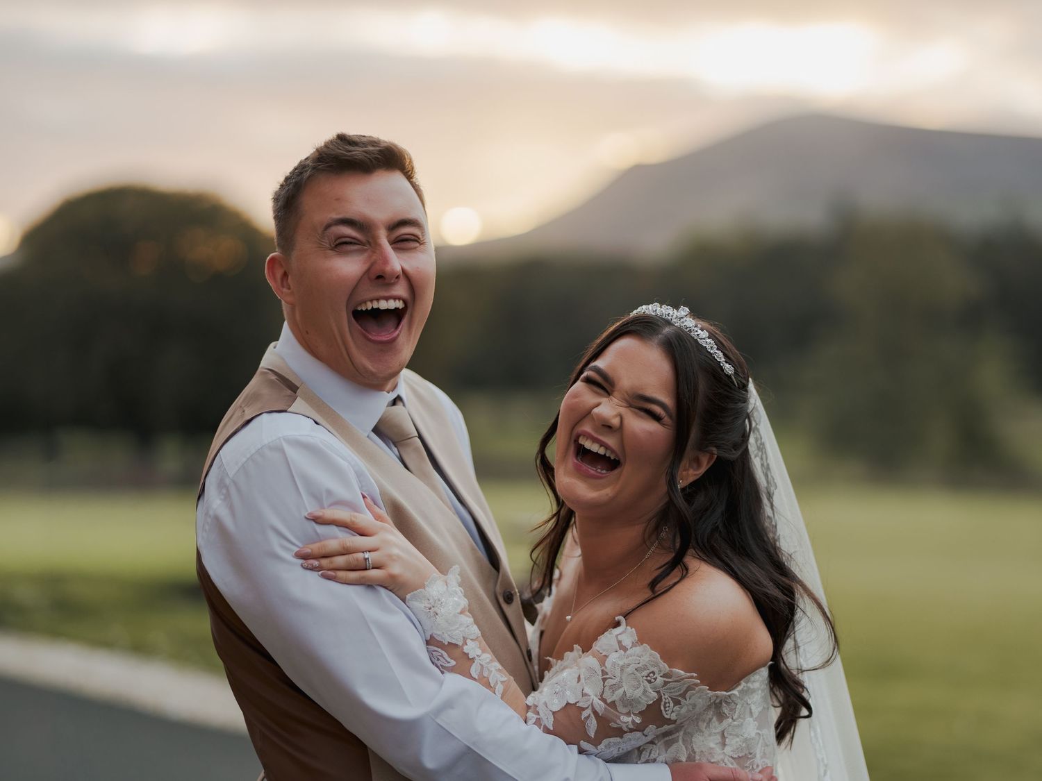Joyful bride and groom laughing together outdoors at sunset with scenic mountain backdrop on their wedding day.