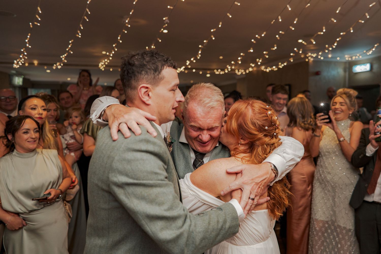 People embrace emotionally during a wedding reception while surrounded by guests and string lights.