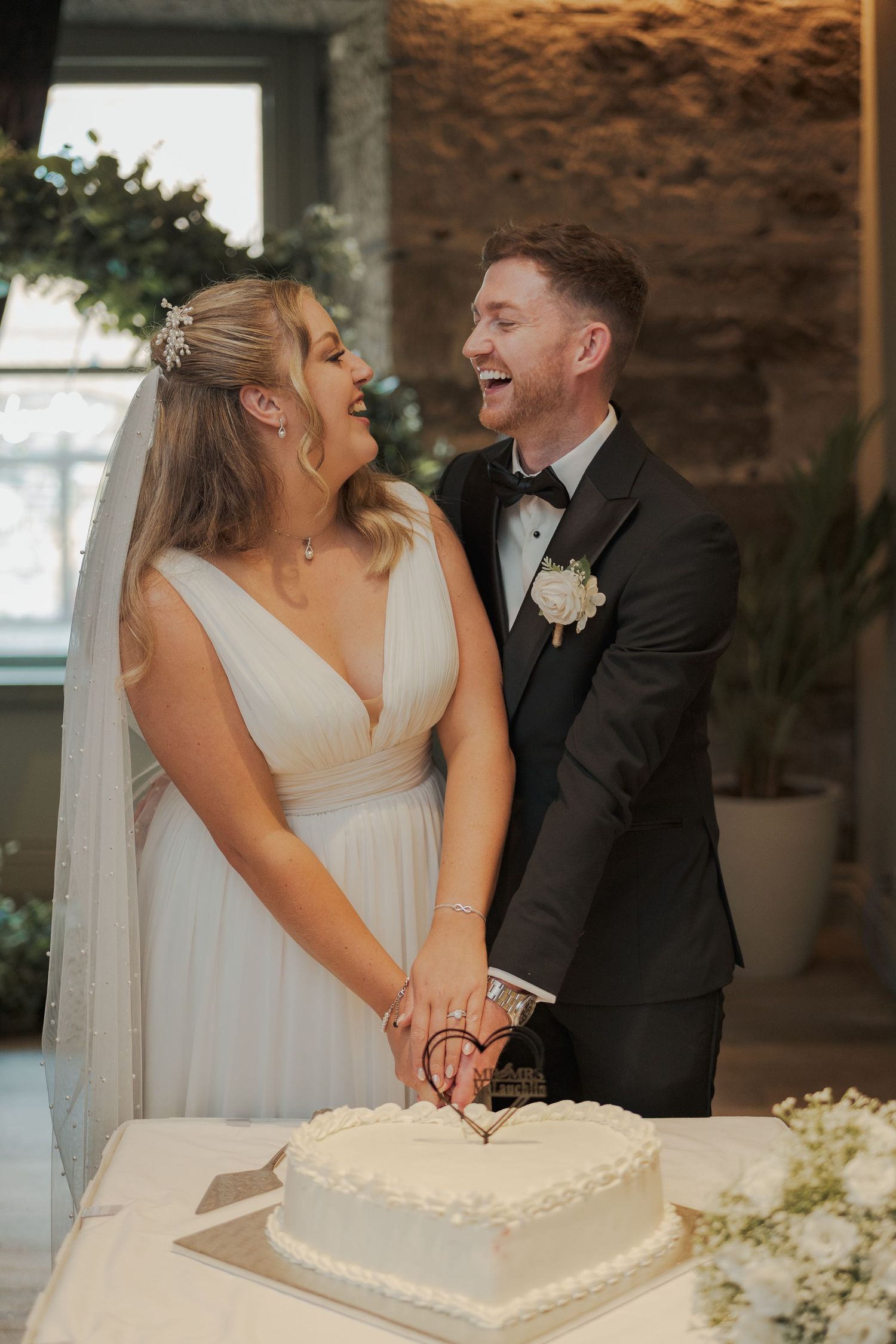 A joyful couple shares a laugh while cutting their heart-shaped white wedding cake together.