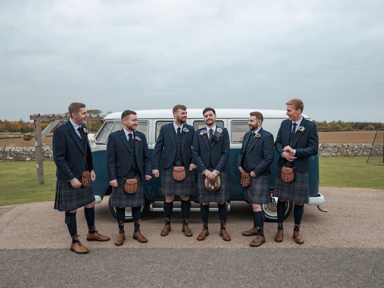 Groomsmen in navy suits sit on a stone wall showing off their brown leather dress shoes and colorful socks.